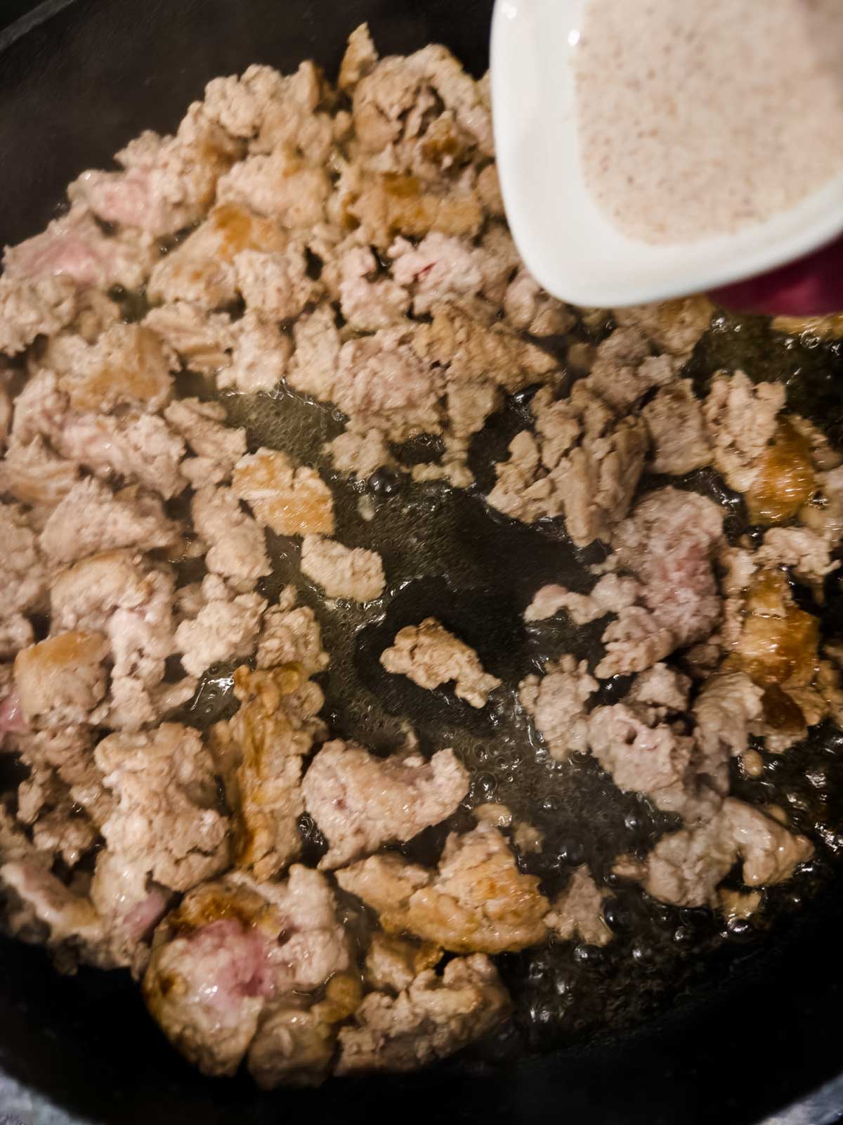 Ground meat is browning in a skillet while a white sauce is being poured in from a small white container.