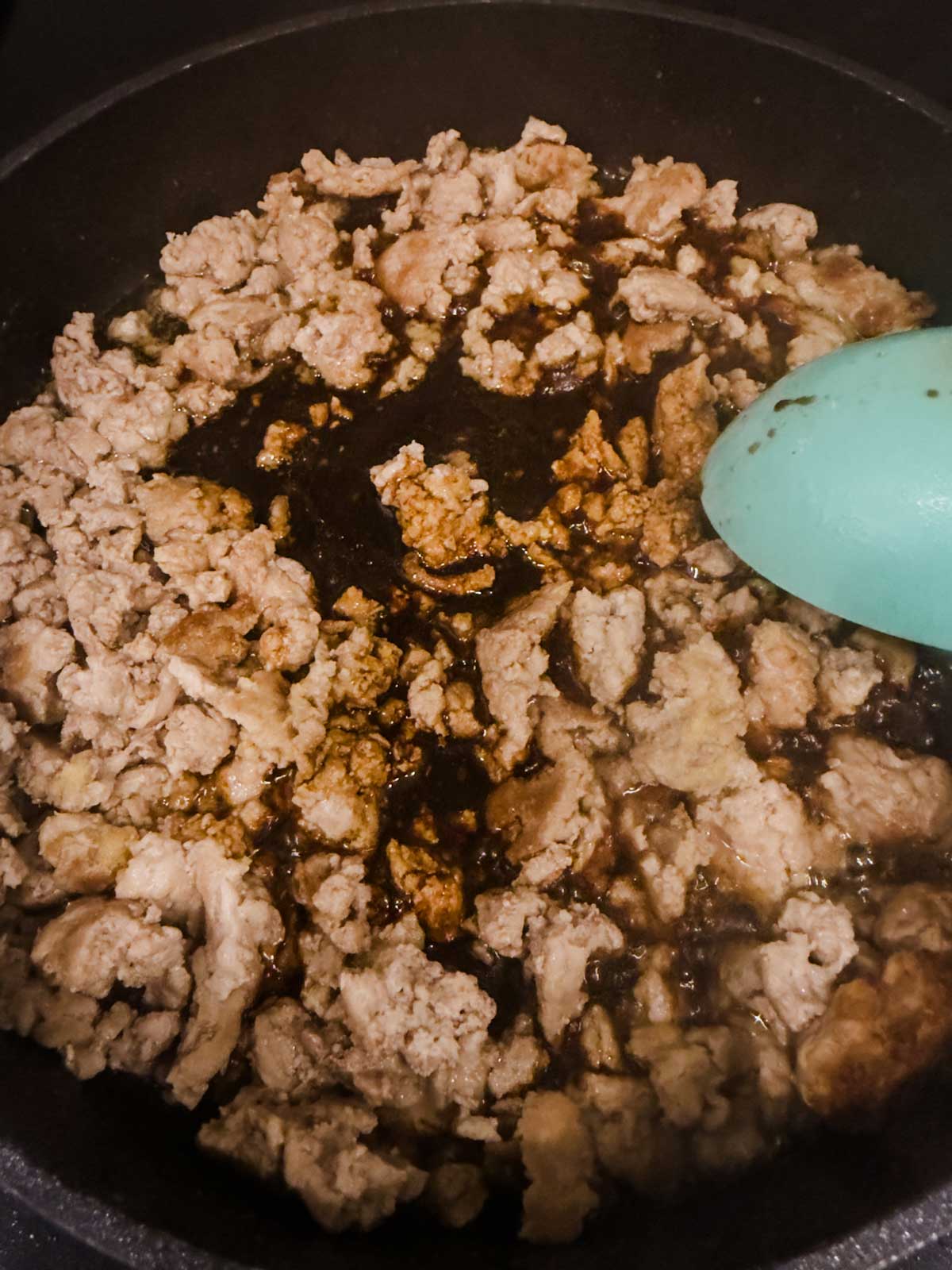 Cooked ground meat in a skillet with a dark sauce in the center, being stirred with a light blue spatula.