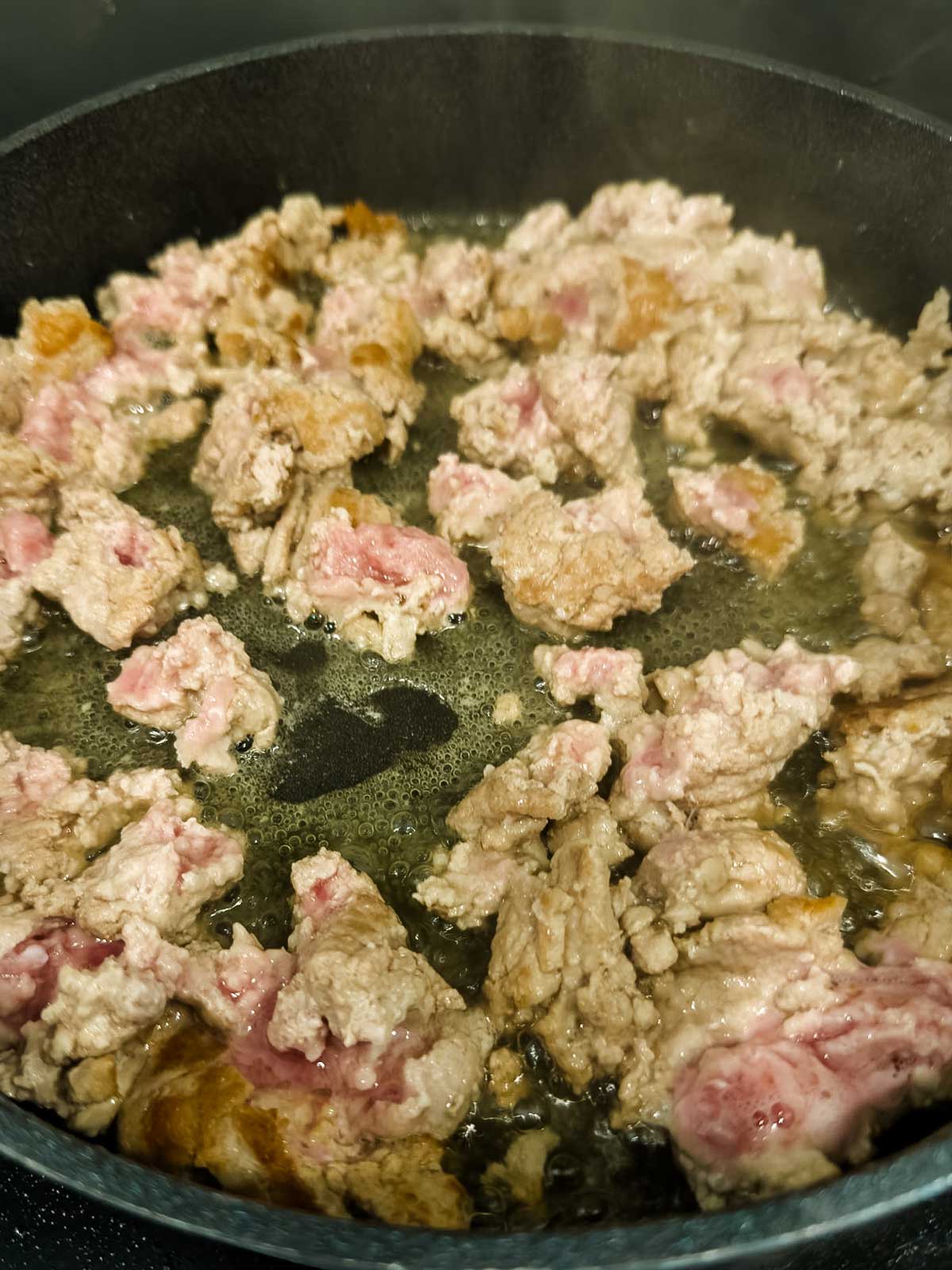 Ground meat is being cooked in a pan. Some pieces are browned while others are still pink, indicating the meat is partially cooked. Oil or juices can be seen bubbling around the meat.