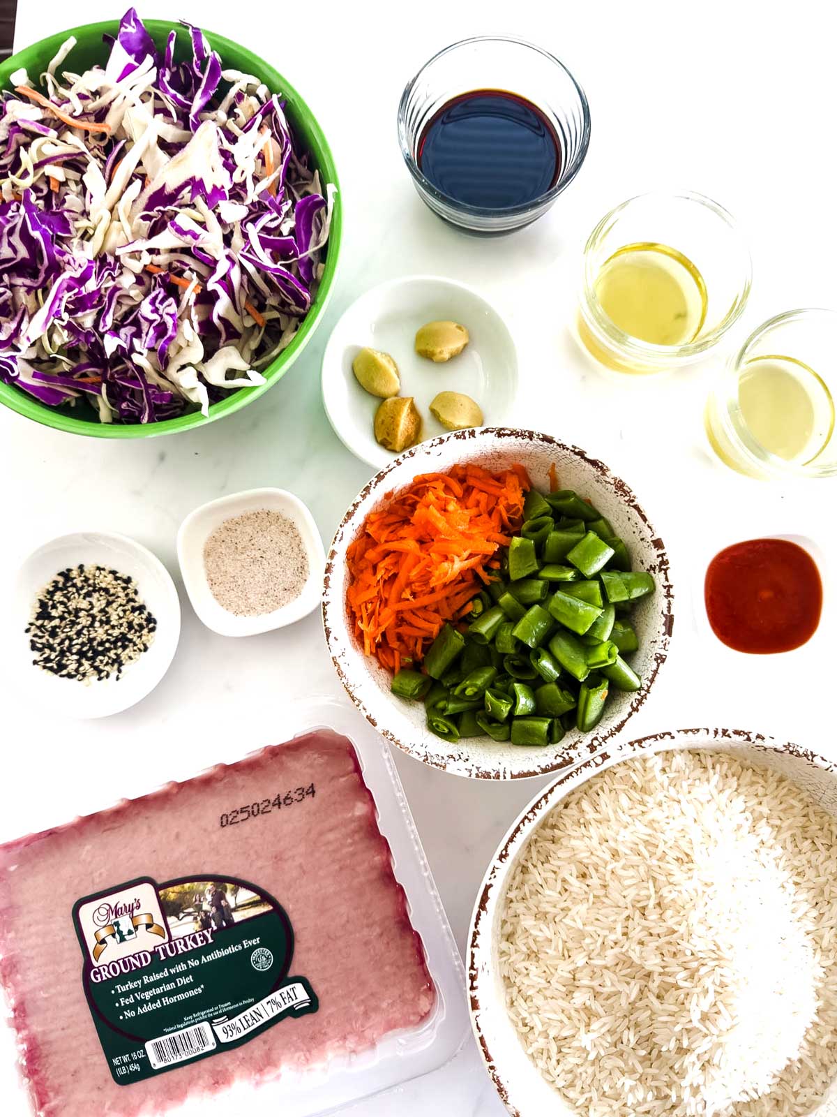 Top-down view of ingredients for an Asian-inspired dish: ground turkey, shredded cabbage, sliced carrots, chopped green bell pepper, rice, garlic cloves, sauces, oil, seasoning, and a bowl of sesame seeds.