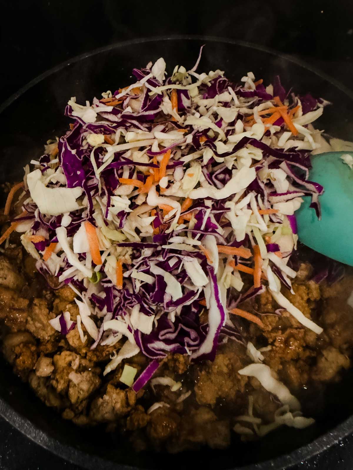 Ground meat cooking in a pan with shredded cabbage, purple cabbage, and carrots on top, being stirred with a teal spatula.