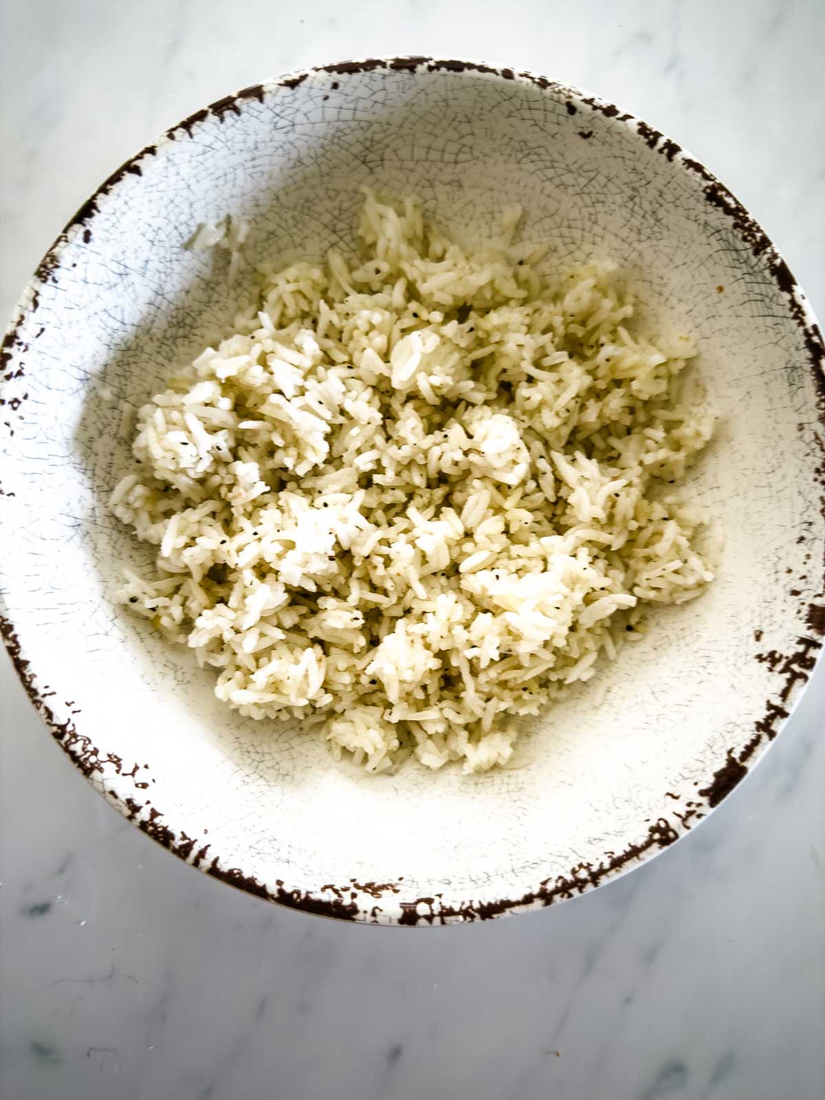 A rustic ceramic bowl filled with cooked white rice sits on a white marble surface. The rice appears fluffy and lightly seasoned.