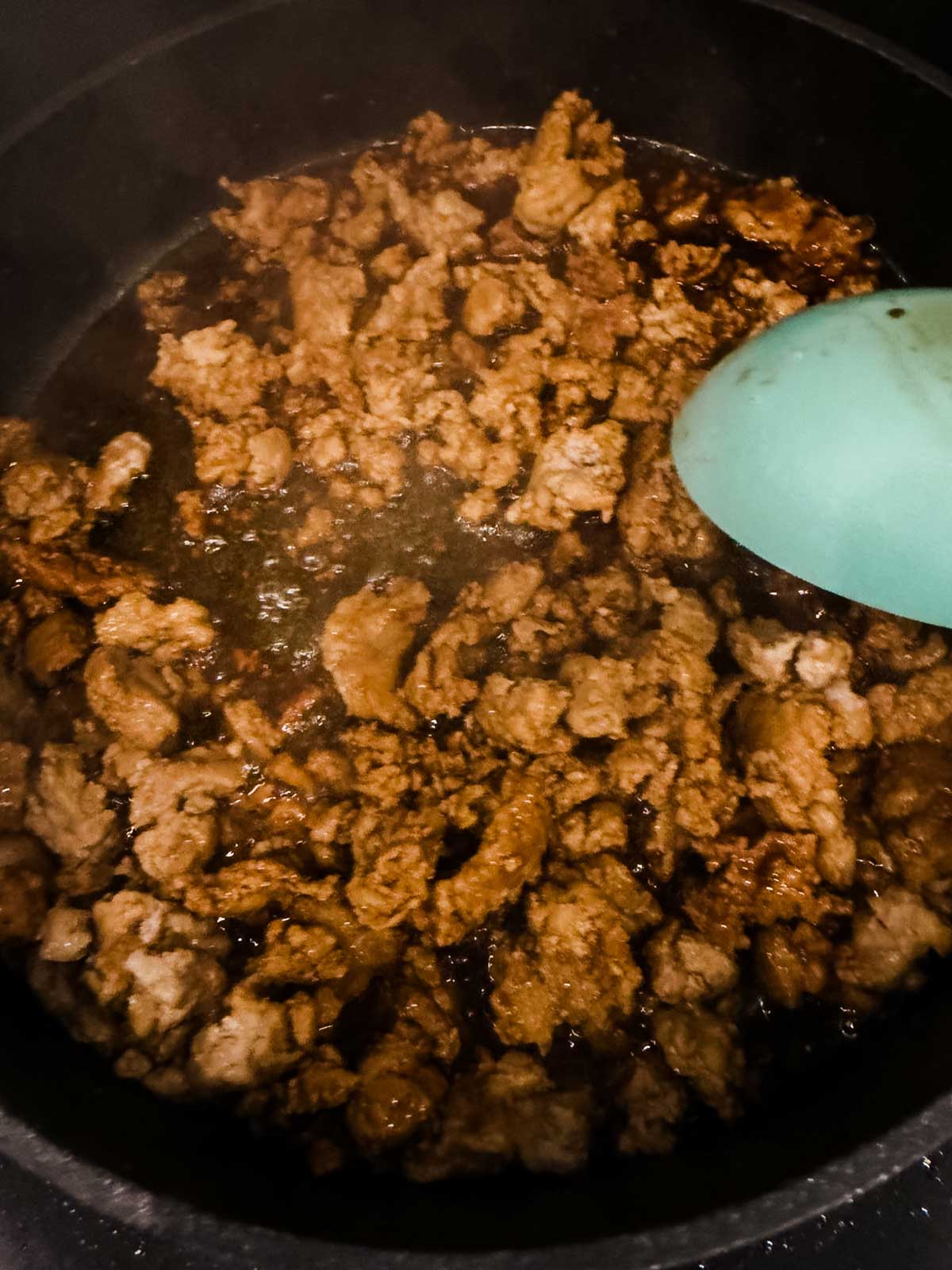 Ground meat browning in a dark skillet, with a blue spatula stirring the mixture. The meat is cooked and slightly crispy around the edges, with some liquid visible at the bottom of the pan.