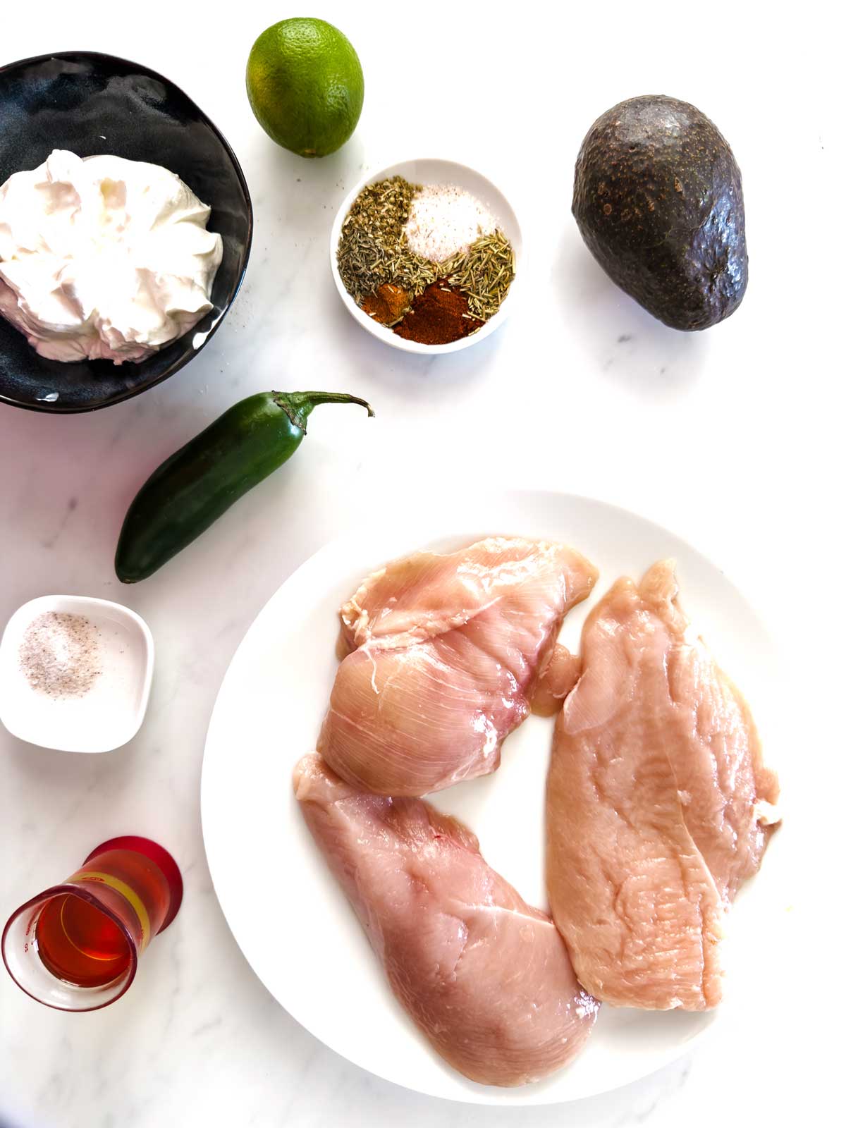 A white surface with raw chicken breasts on a plate, surrounded by a lime, avocado, jalapeño, bowl of yogurt, mixed spices in a small dish, a salt container, and a small glass of red liquid.
