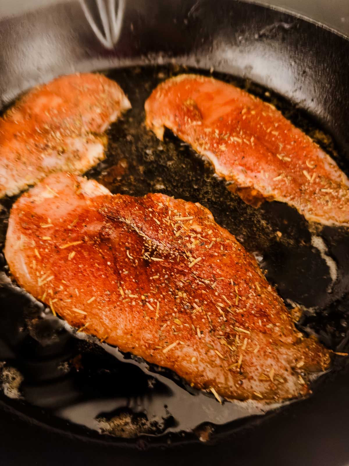 Three seasoned chicken breasts are sizzling in a black cast iron skillet, surrounded by oil. The meat is coated with various spices and herbs, giving it a rich, flavorful appearance.