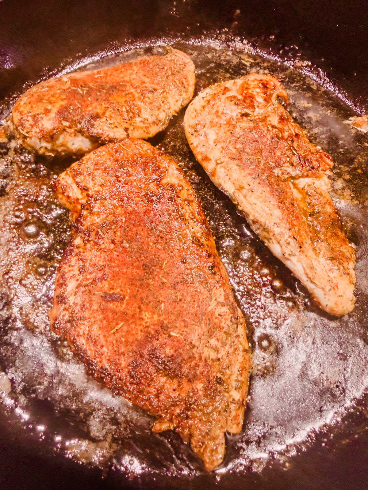 Three seasoned chicken breasts are sizzling in a black skillet, surrounded by bubbling oil and cooking juices. The chicken has a browned, crispy exterior.