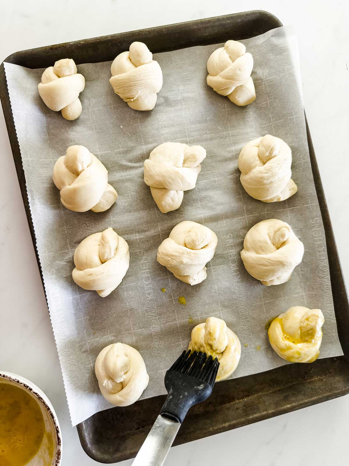 A baking tray lined with parchment paper holds twelve unbaked, twisted dough knots. A pastry brush is applying an egg wash to one knot, with a bowl of egg wash visible in the lower left corner.