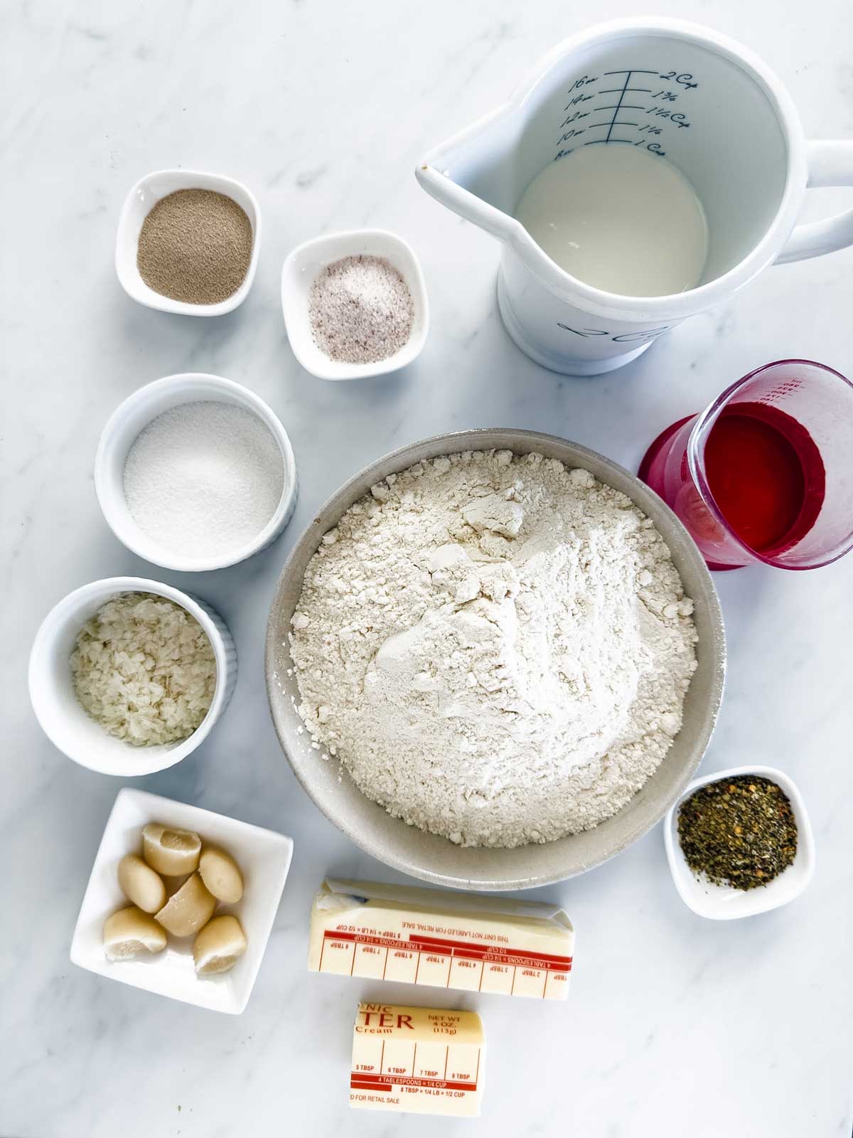 A variety of ingredients in separate bowls and containers on a white surface, including flour, milk, beet juice, dry yeast, sugar, salt, chopped onions, garlic cloves, two sticks of butter, and a small bowl of herbs.