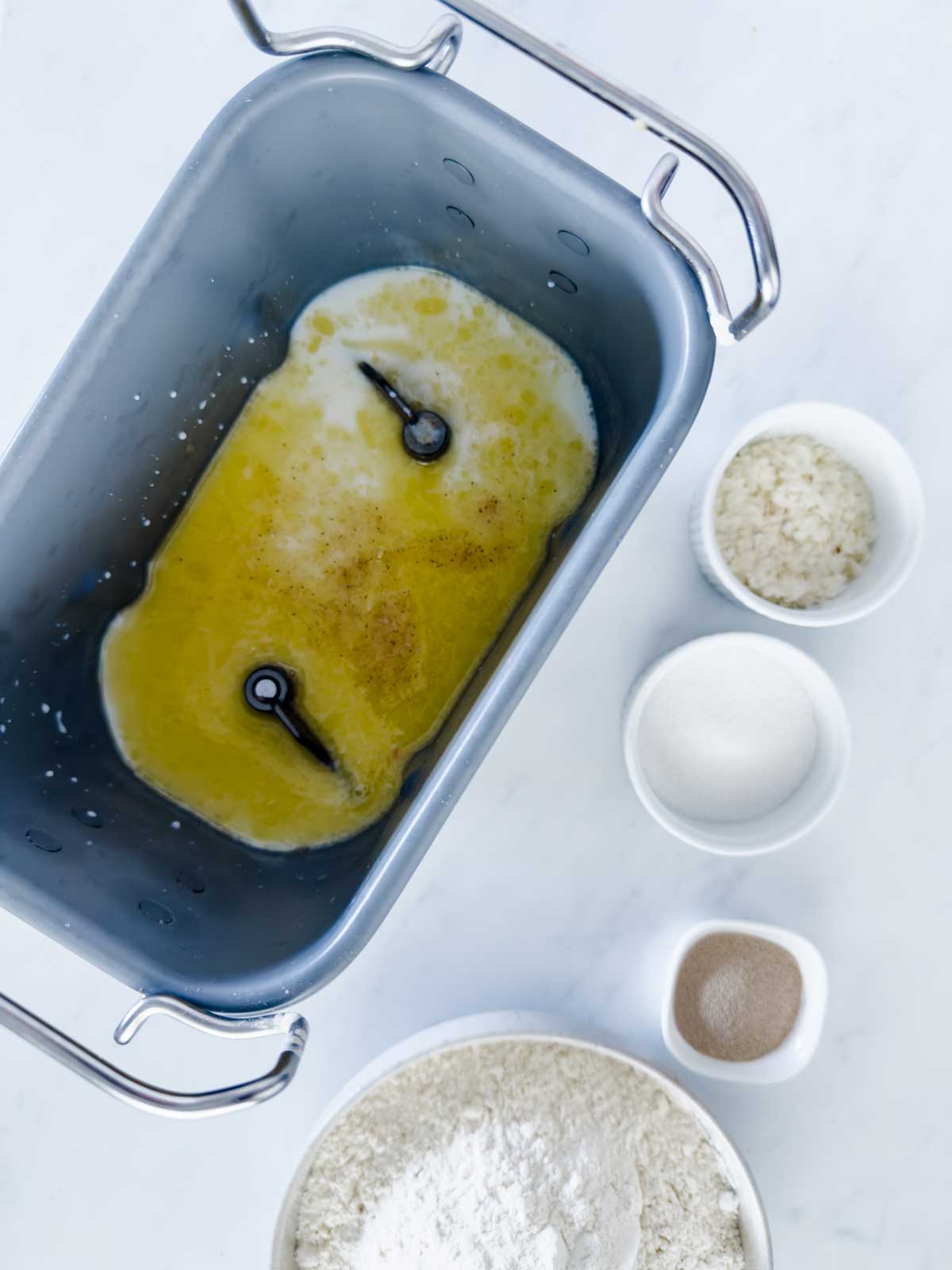 A bread machine pan with melted butter and liquid next to small bowls containing potato flakes, sugar, yeast, and a larger bowl of flour on a white surface.