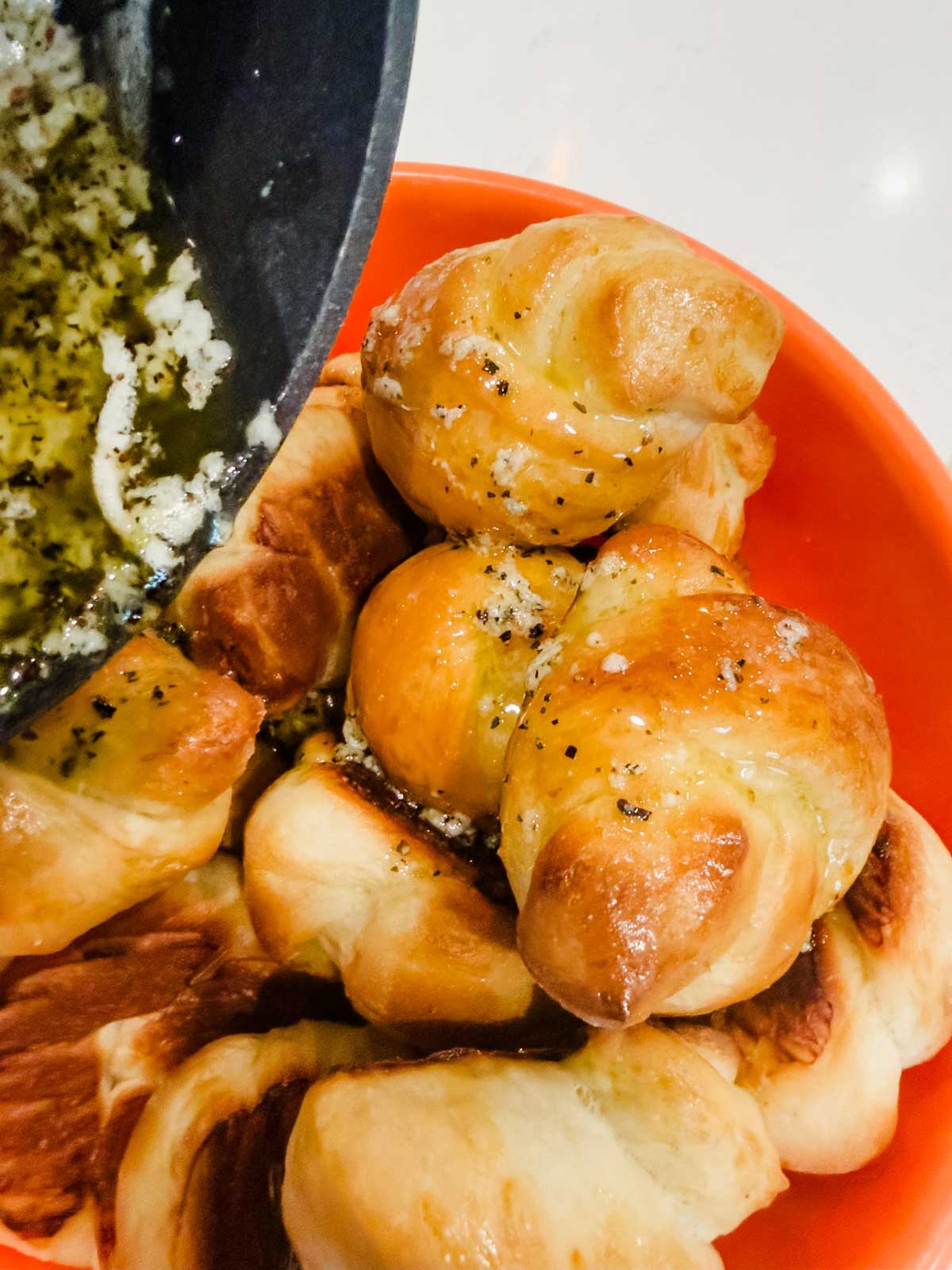 A close-up of golden brown, twisted bread rolls in an orange bowl, being drizzled with a mixture of melted butter, garlic, and herbs from a pan.