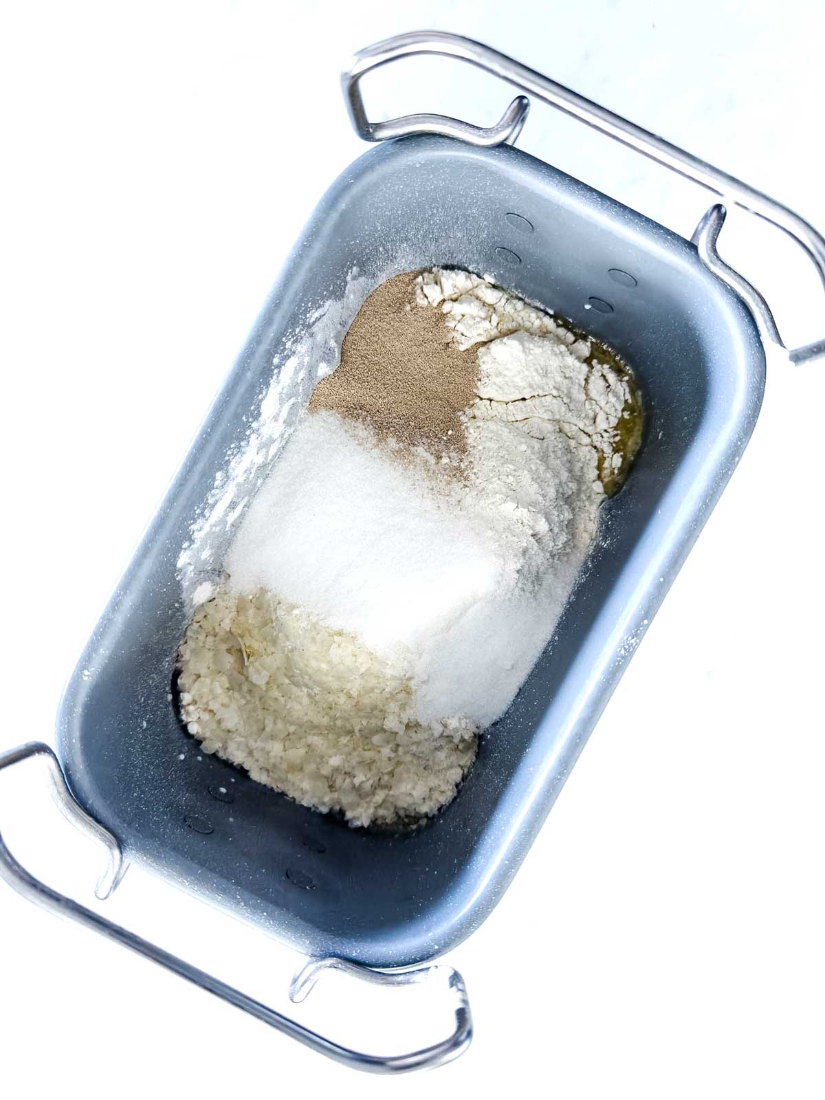 A bread machine pan containing separated ingredients: flour, sugar, yeast, and rice, viewed from above on a white background.