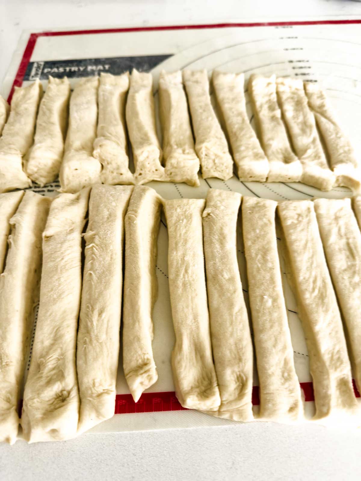 Unbaked bread dough strips are arranged in rows on a pastry mat, ready to be baked.