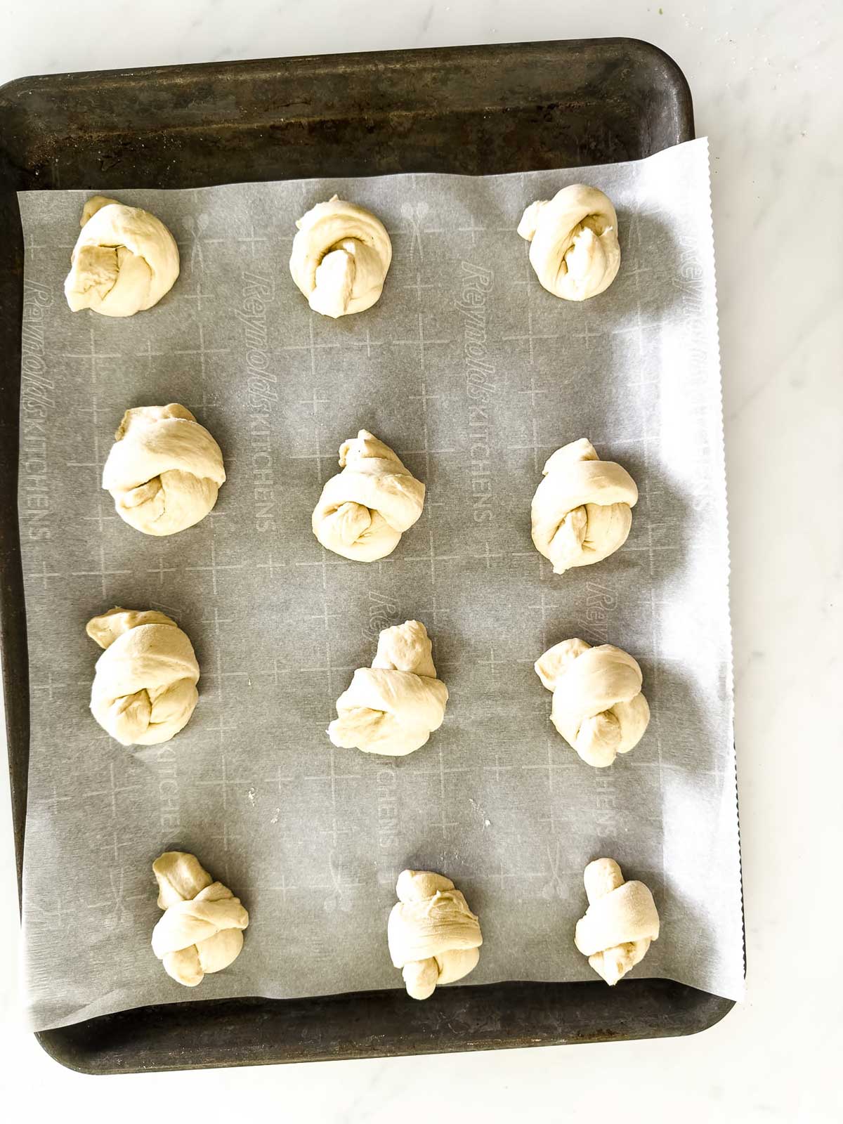 Twelve pieces of knotted dough are evenly spaced on a parchment-lined baking sheet, ready to be baked. The tray sits on a white surface.