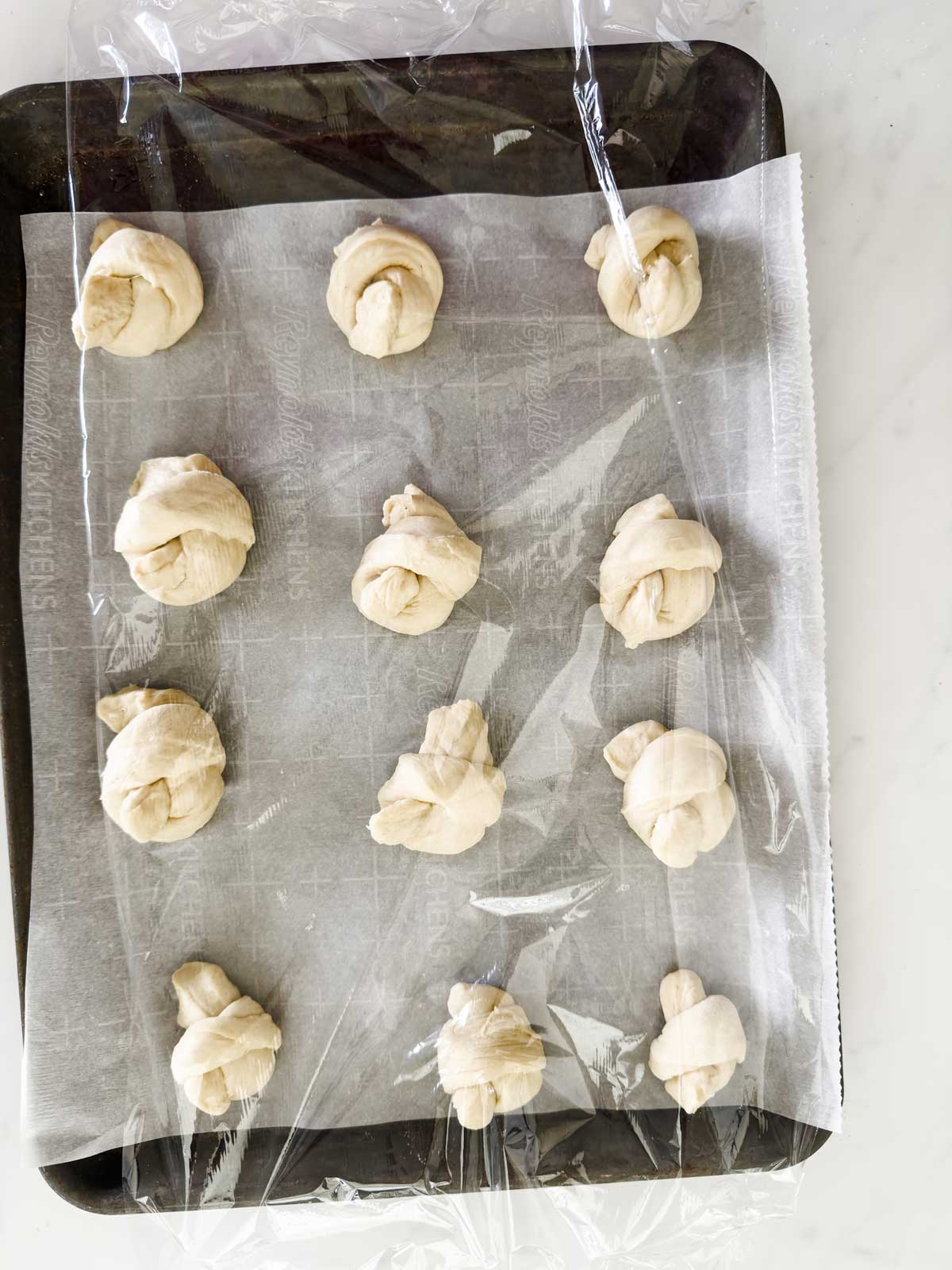 Twelve unbaked dough knots are arranged on a parchment-lined baking sheet. The sheet is covered with a layer of plastic wrap, and everything is set on a white surface.