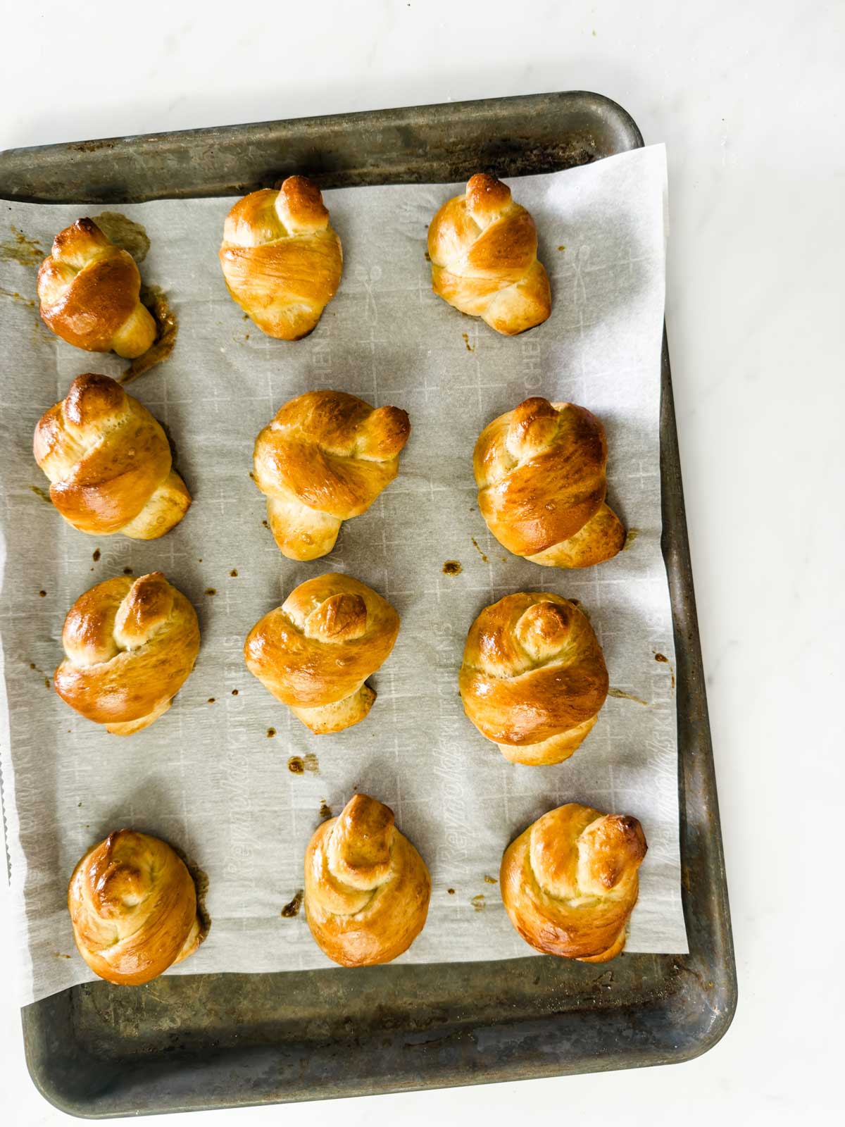 A baking sheet lined with parchment paper holds twelve golden-brown, twisted bread rolls arranged in rows on a white surface.