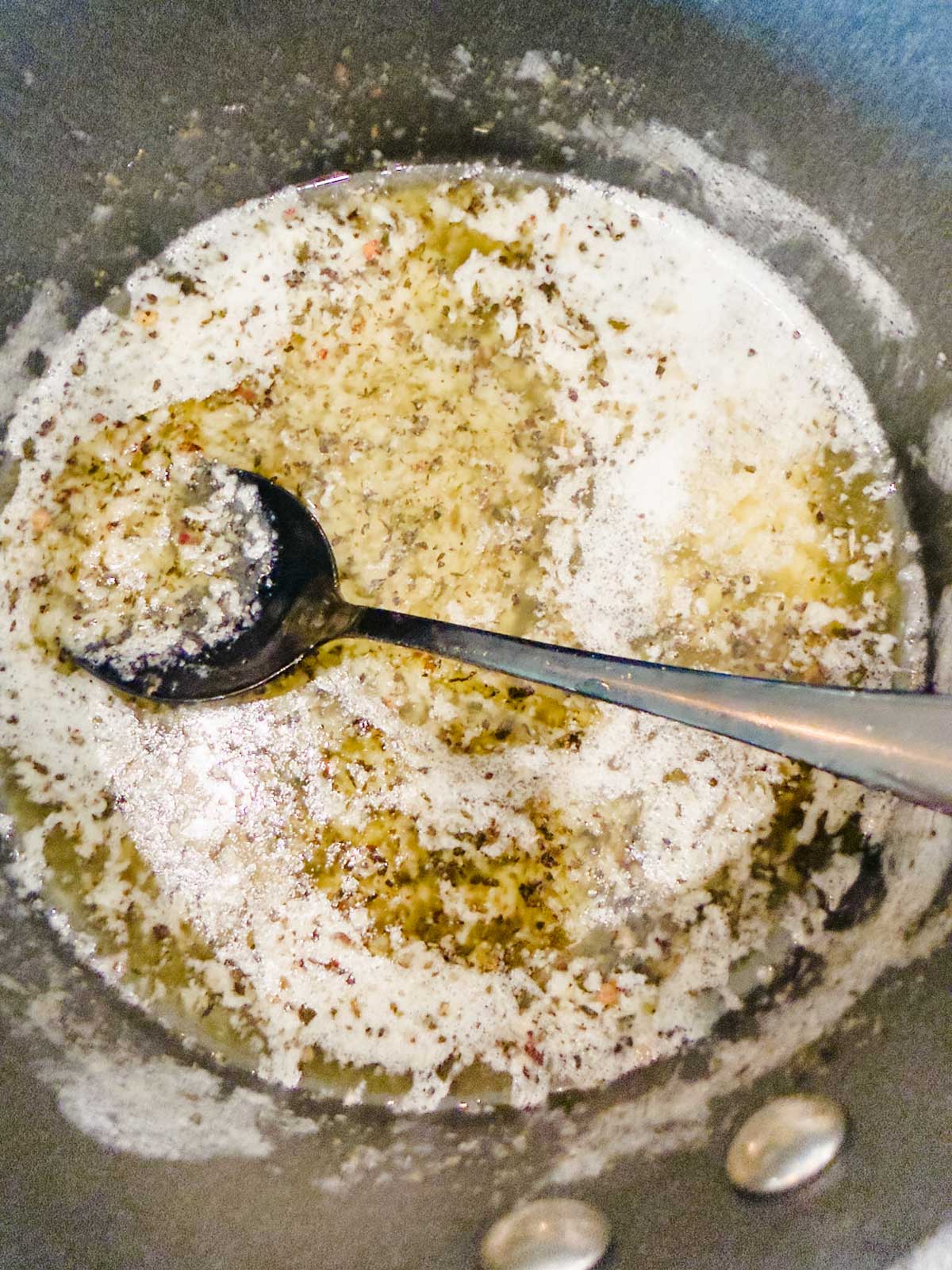 A metal spoon rests in a saucepan with melted butter, herbs, and garlic being sautéed, creating a bubbly mixture.