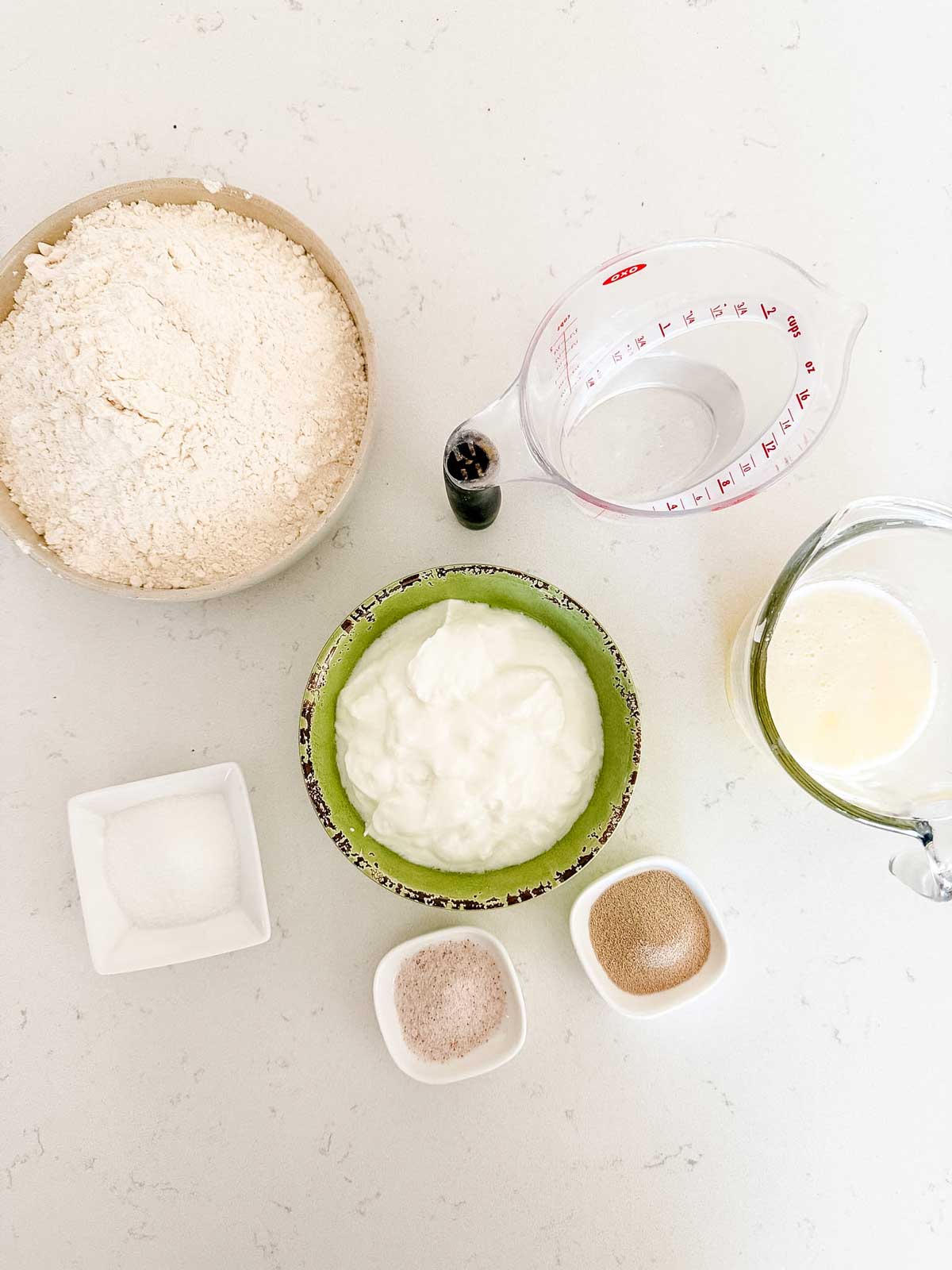 Overhead view of baking ingredients: a bowl of flour, a measuring cup of water, a bowl of yogurt, a glass of melted butter, a small dish of salt, and two small dishes with yeast and sugar on a white surface.