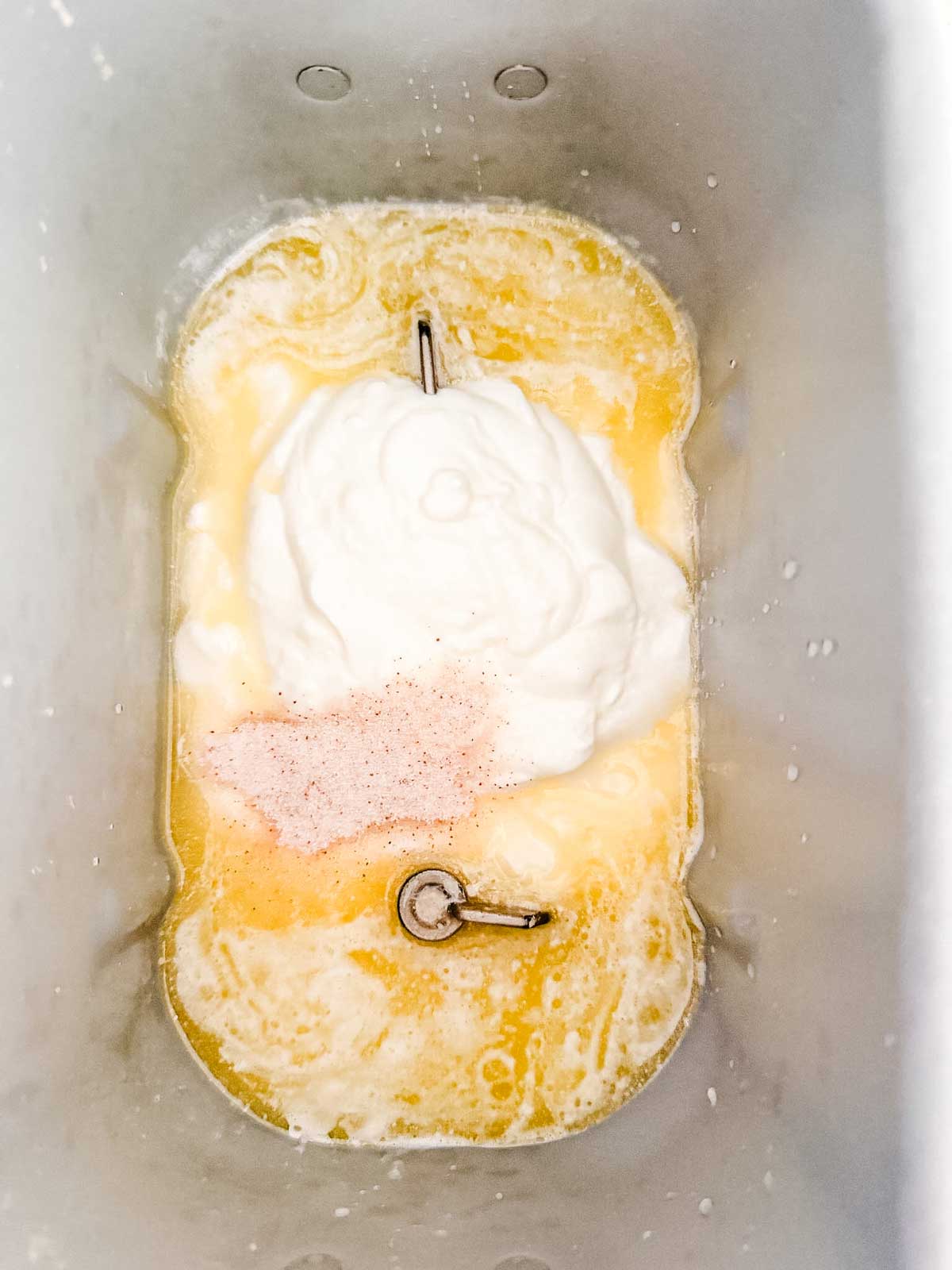 Overhead view of ingredients in a bread machine pan, including milk, water, yogurt, melted butter, and a sprinkle of salt, ready to be mixed for baking.