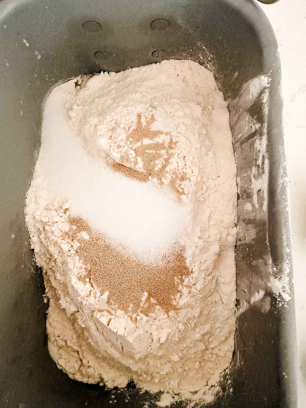 A bread machine pan filled with flour, granulated sugar, and dry yeast, each ingredient forming separate piles on top of the flour.