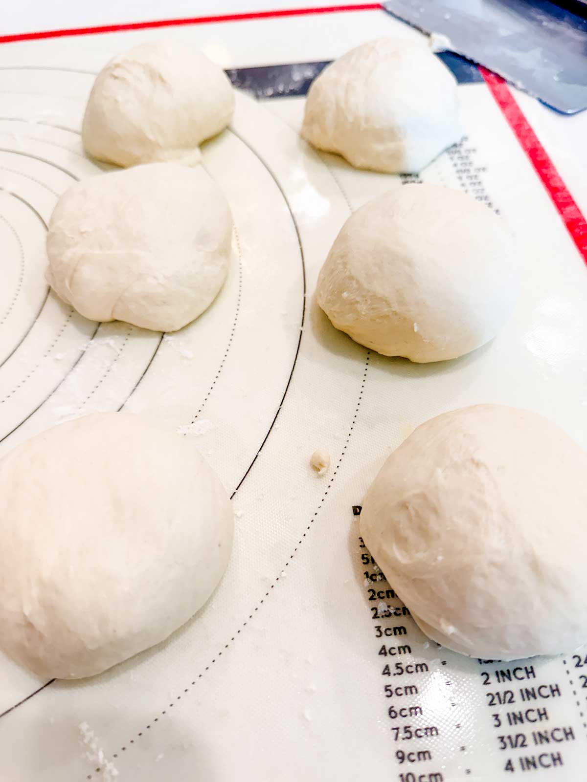Six balls of dough rest on a silicone baking mat with measurement markings in inches and centimeters, ready for baking or further preparation.