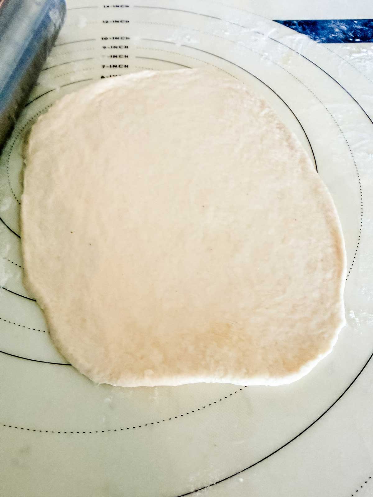 A sheet of dough rolled out on a circular measurement mat, with a rolling pin partially visible in the upper left corner.