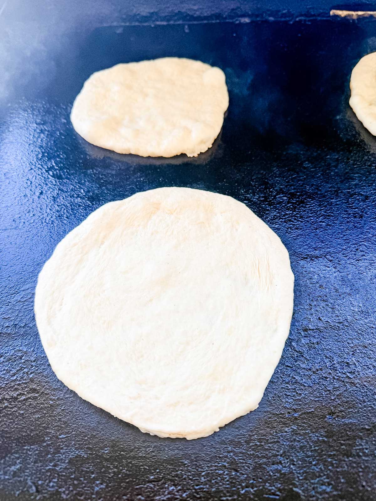 Three raw, round pieces of dough are cooking on a dark, flat Blackstone griddle surface, ready to be made into flatbreads or tortillas. Steam or heat is visible around the dough.