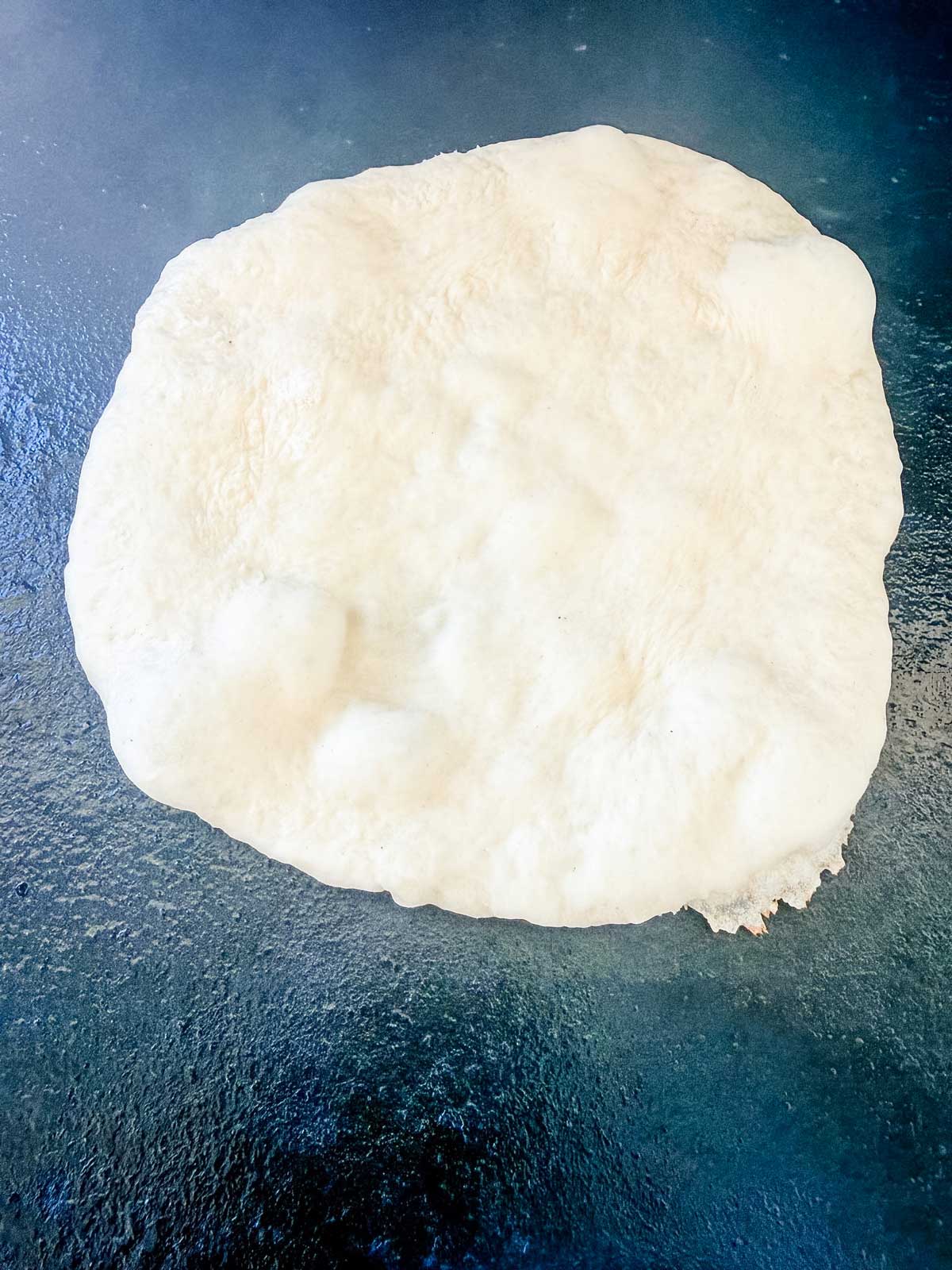 A round, uncooked naan dough rests on a dark, textured surface, showing a few air bubbles and an uneven edge.