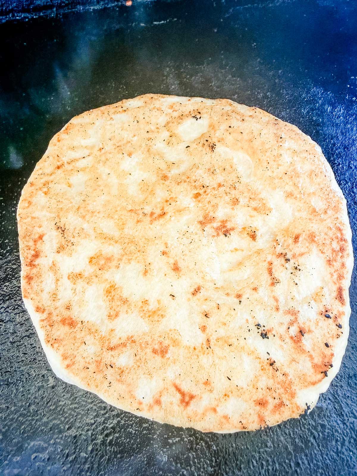 A round, golden-brown flatbread is cooking on a dark griddle, with visible spots of browning and a slightly textured surface.