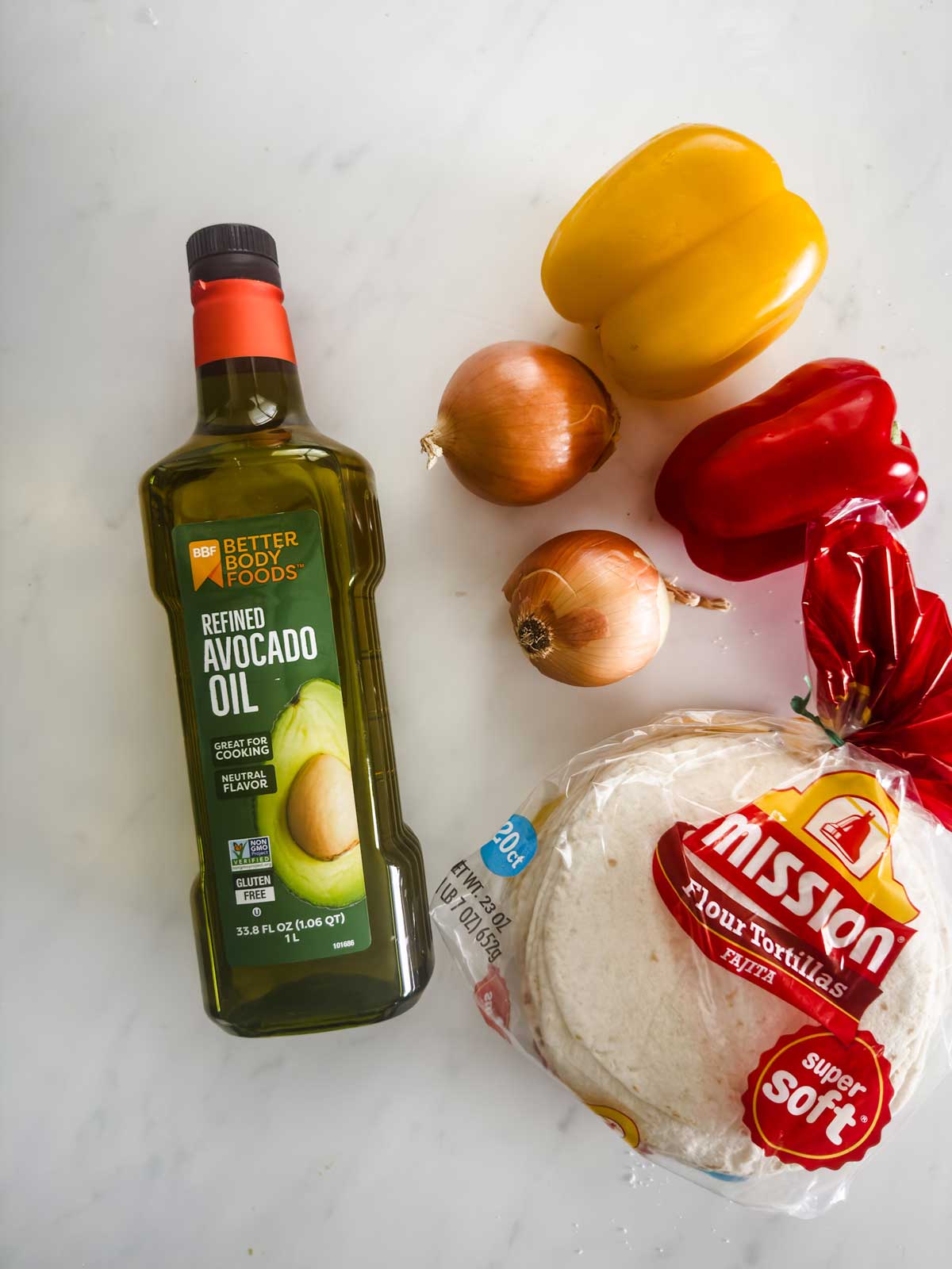 A bottle of refined avocado oil, two yellow onions, a yellow bell pepper, a red bell pepper, and a package of Mission flour tortillas on a white surface.