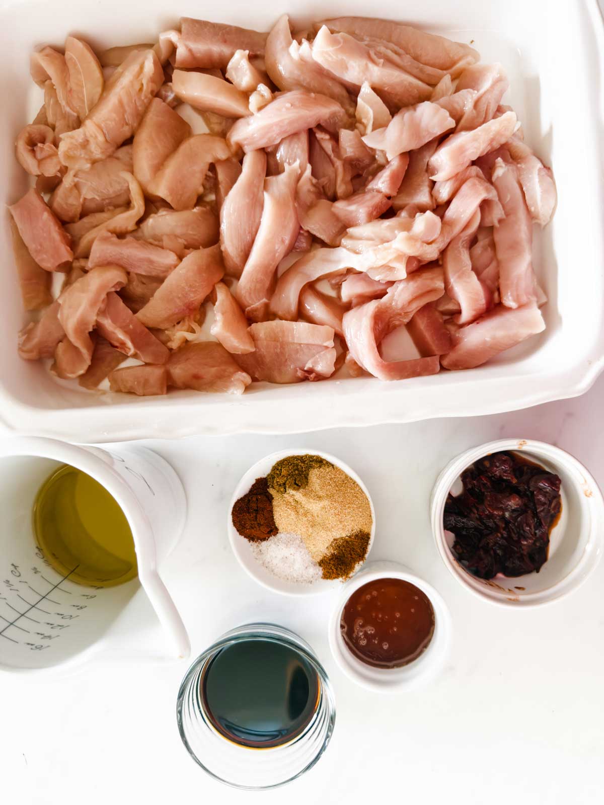 Raw sliced chicken in a white dish, surrounded by small bowls containing oil, spices, dried chilies, sauce, and a cup of dark liquid on a white surface.