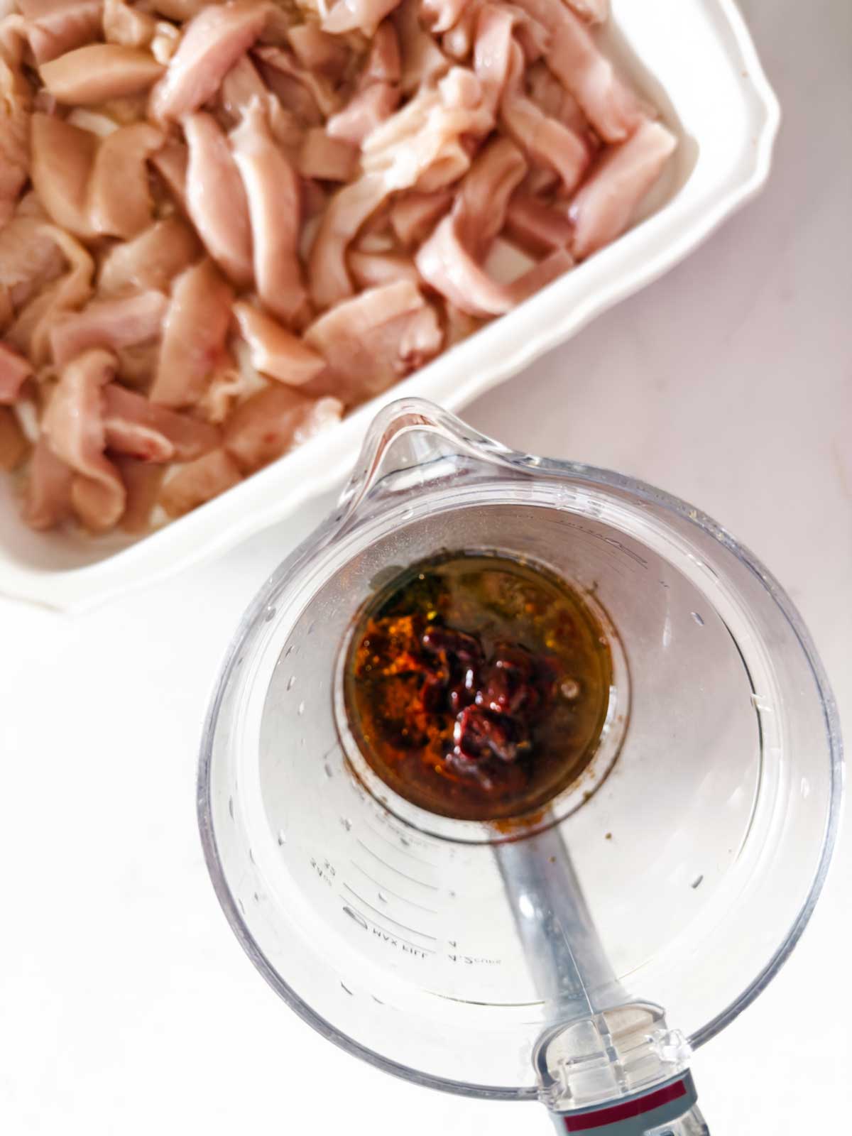Top view of a blender with sauce ingredients, next to a dish filled with raw, cut chicken pieces on a white surface.