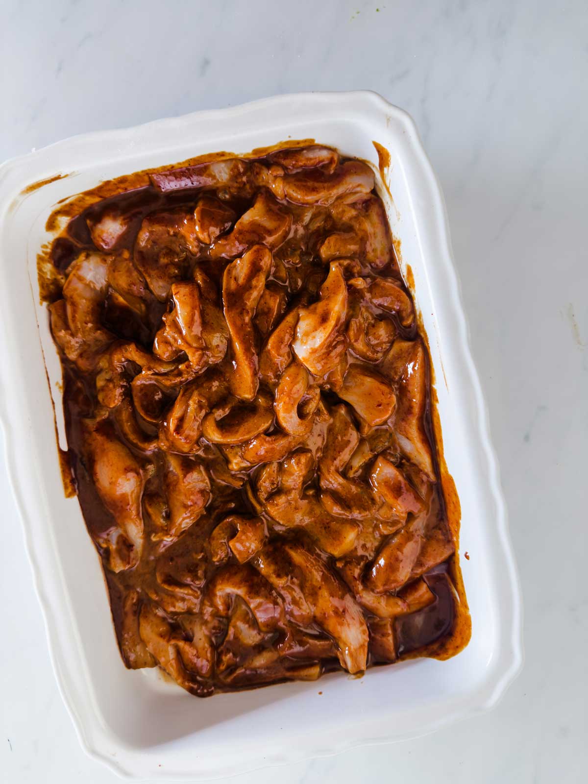 Raw, sliced chicken marinated in a reddish-brown sauce, arranged in a white rectangular dish on a white marble surface.