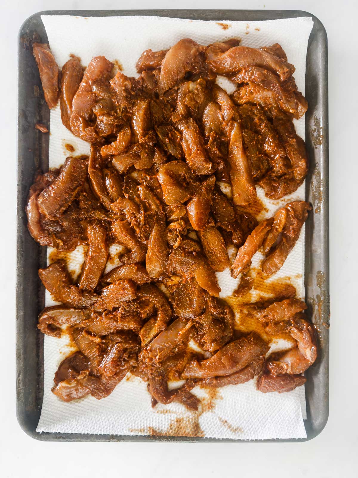 Raw marinated slices of meat spread out on a paper towel-lined baking sheet, ready for cooking. The meat has a reddish-brown seasoning and is arranged in an even layer.