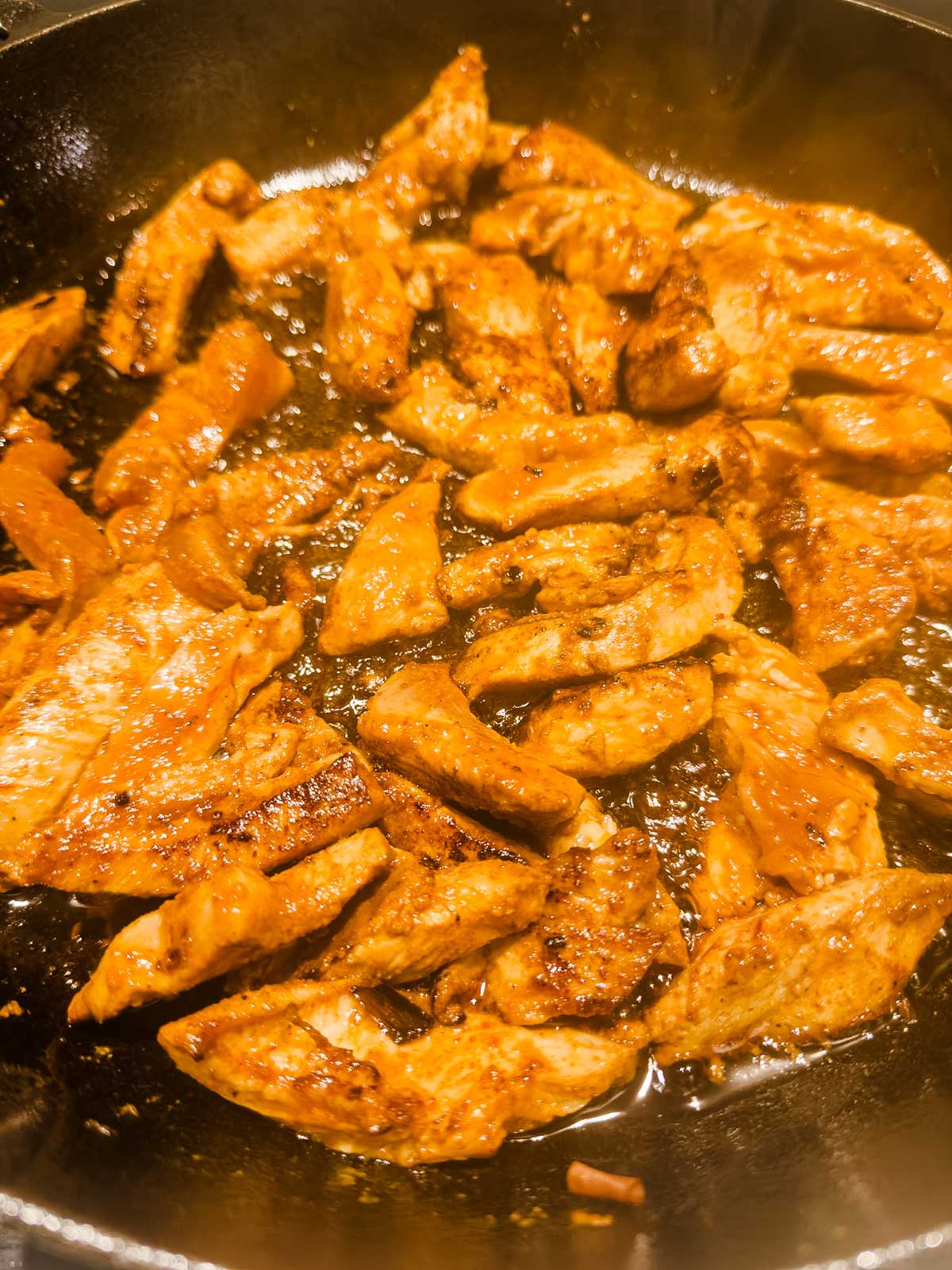 Sliced pieces of seasoned chicken cooking and sizzling in a black skillet, coated with a golden-brown sauce.