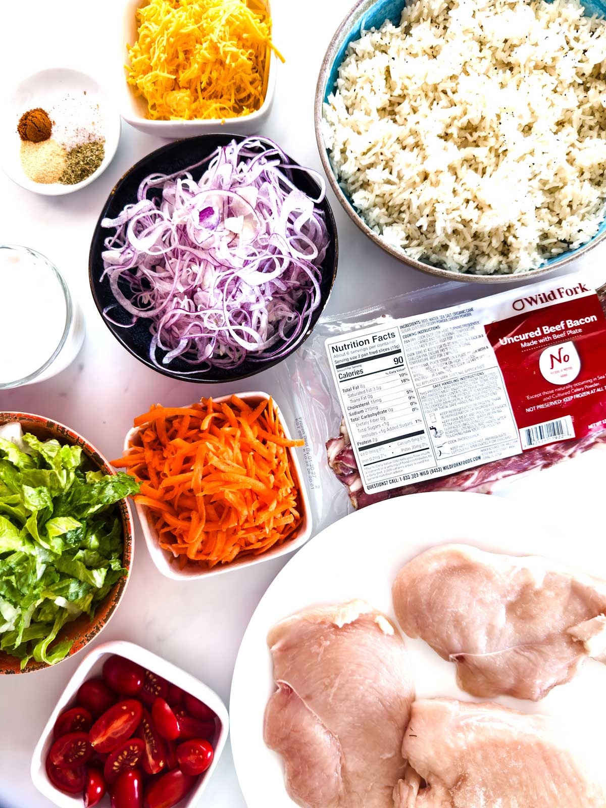Bowls of shredded cheese, sliced red onions, cooked rice, shredded carrots, chopped lettuce, and grape tomatoes sit on a white surface with raw chicken breasts, a package of uncured beef bacon, and a small cup of spices and milk.