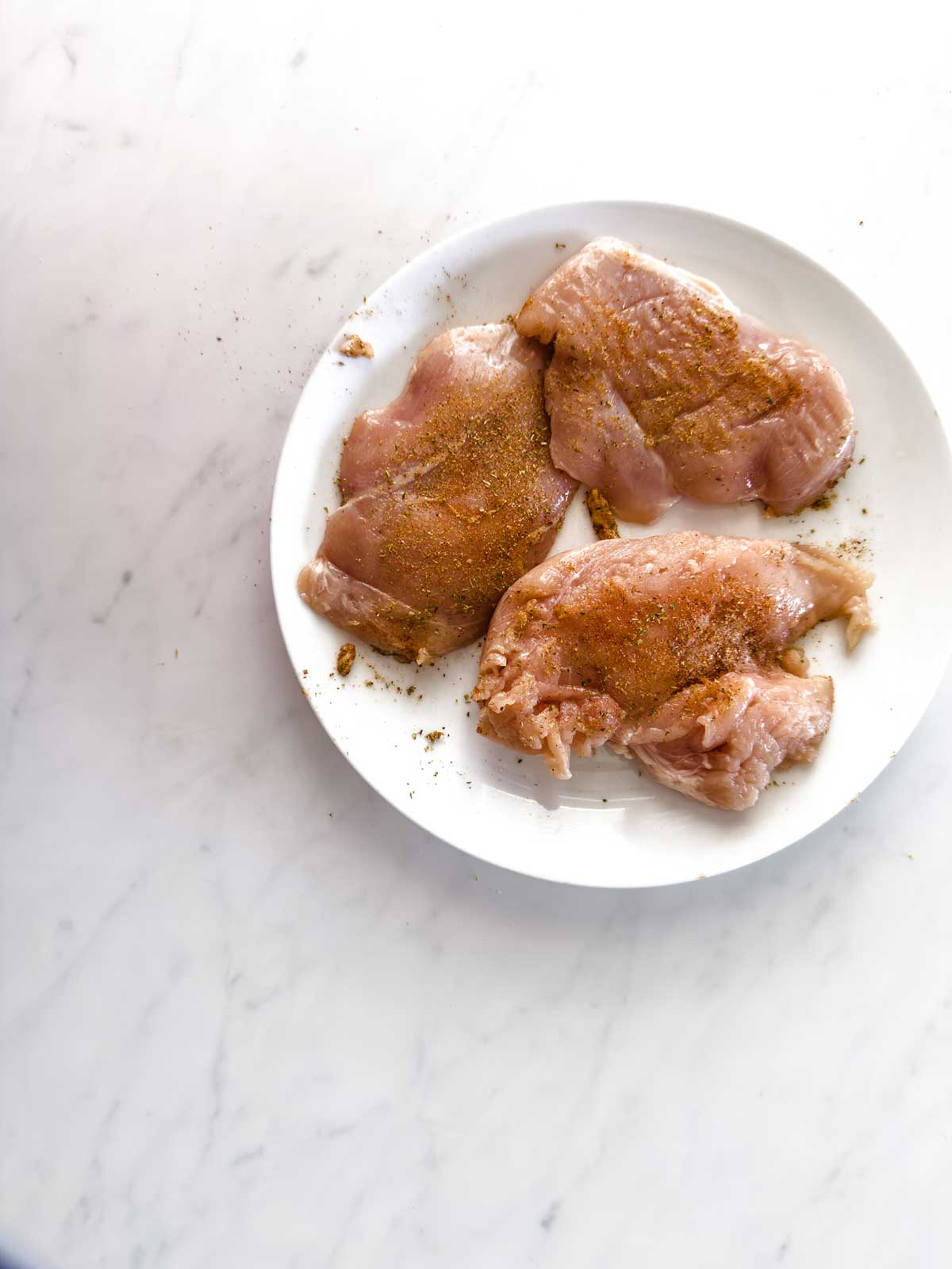 Three raw, seasoned chicken breasts are arranged on a white plate atop a white marble surface. Some of the seasoning has spilled onto the plate.