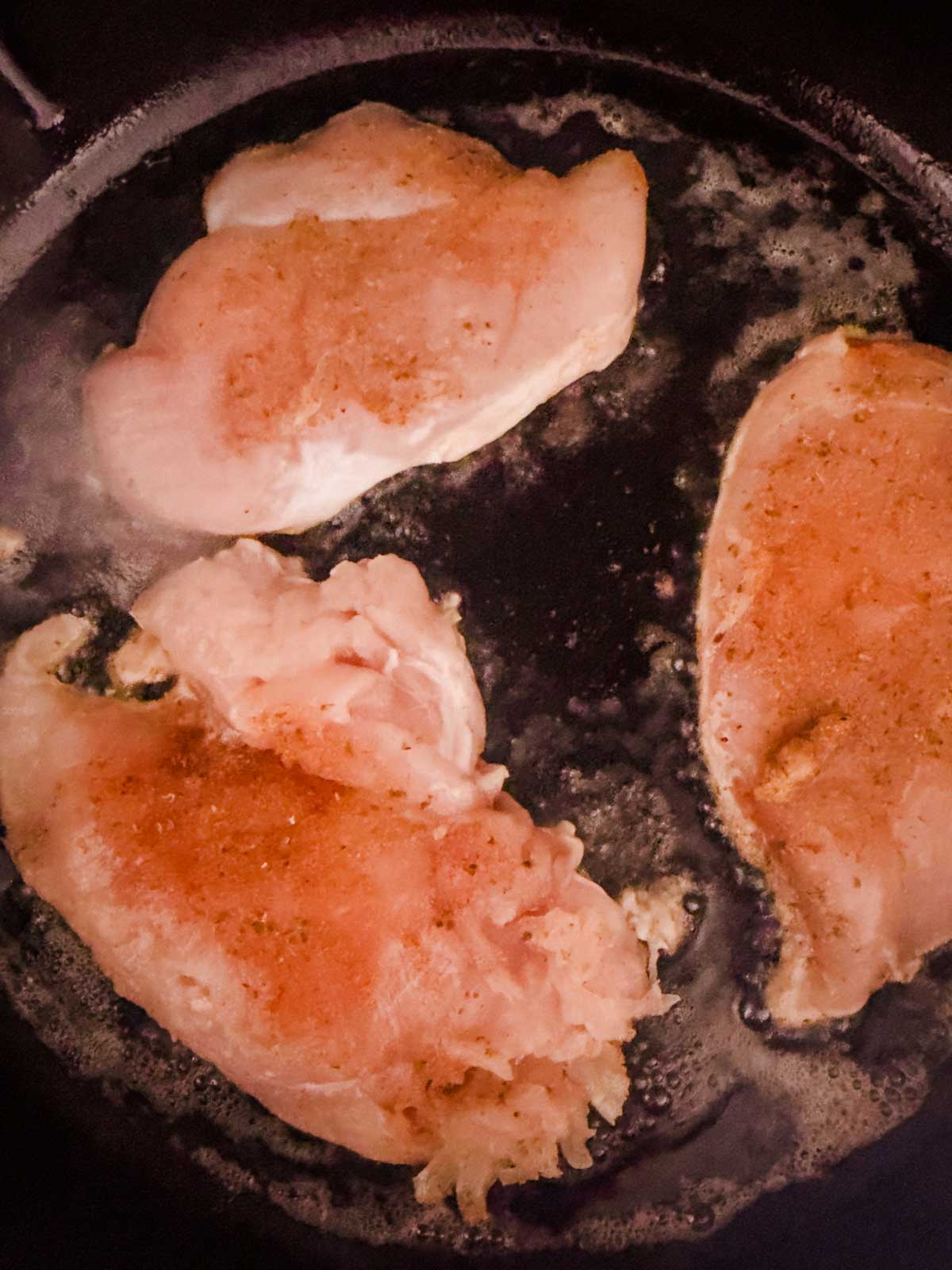 Raw, seasoned chicken breasts cooking in a black skillet with oil, starting to brown around the edges. Steam rises as the chicken cooks.