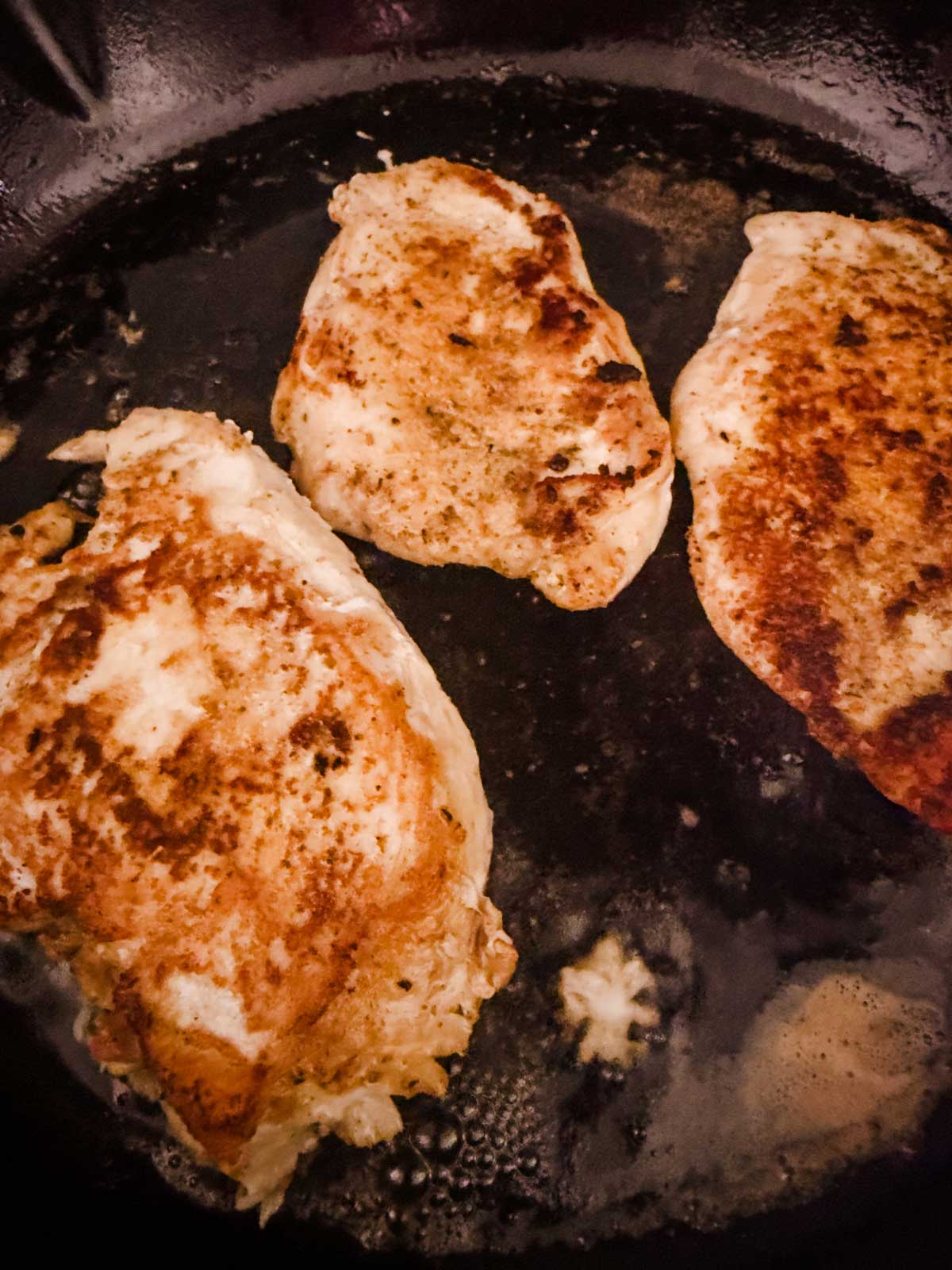 Three pieces of seasoned chicken breast cooking in a dark, oily skillet, with golden-brown sear marks visible on the surface of the meat.