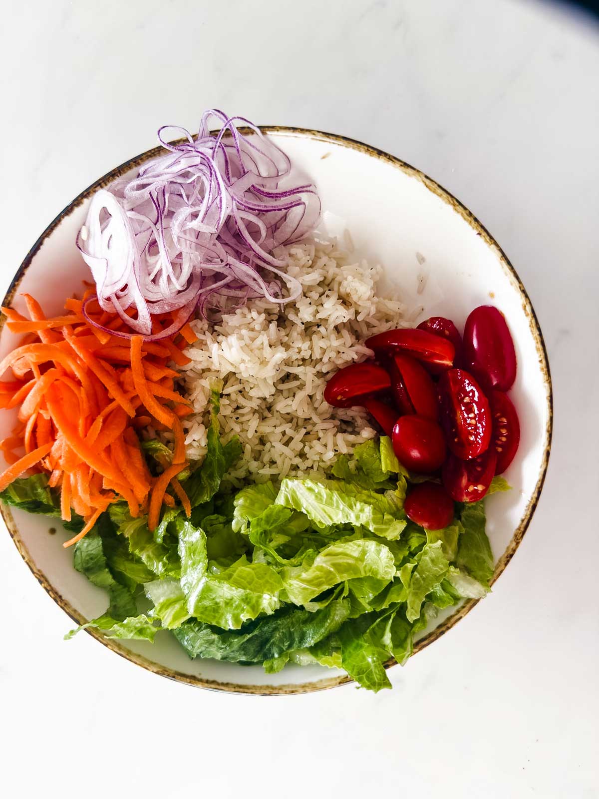 A bowl containing shredded lettuce, sliced cherry tomatoes, cooked rice, shredded carrots, and thinly sliced red onions, arranged in separate sections on a white surface.