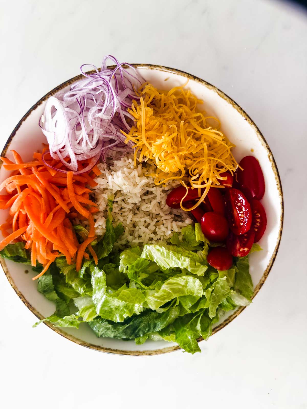 A bowl filled with chopped lettuce, shredded carrots, sliced red onions, shredded cheddar cheese, grape tomatoes, and a serving of rice, arranged in sections on a white surface.