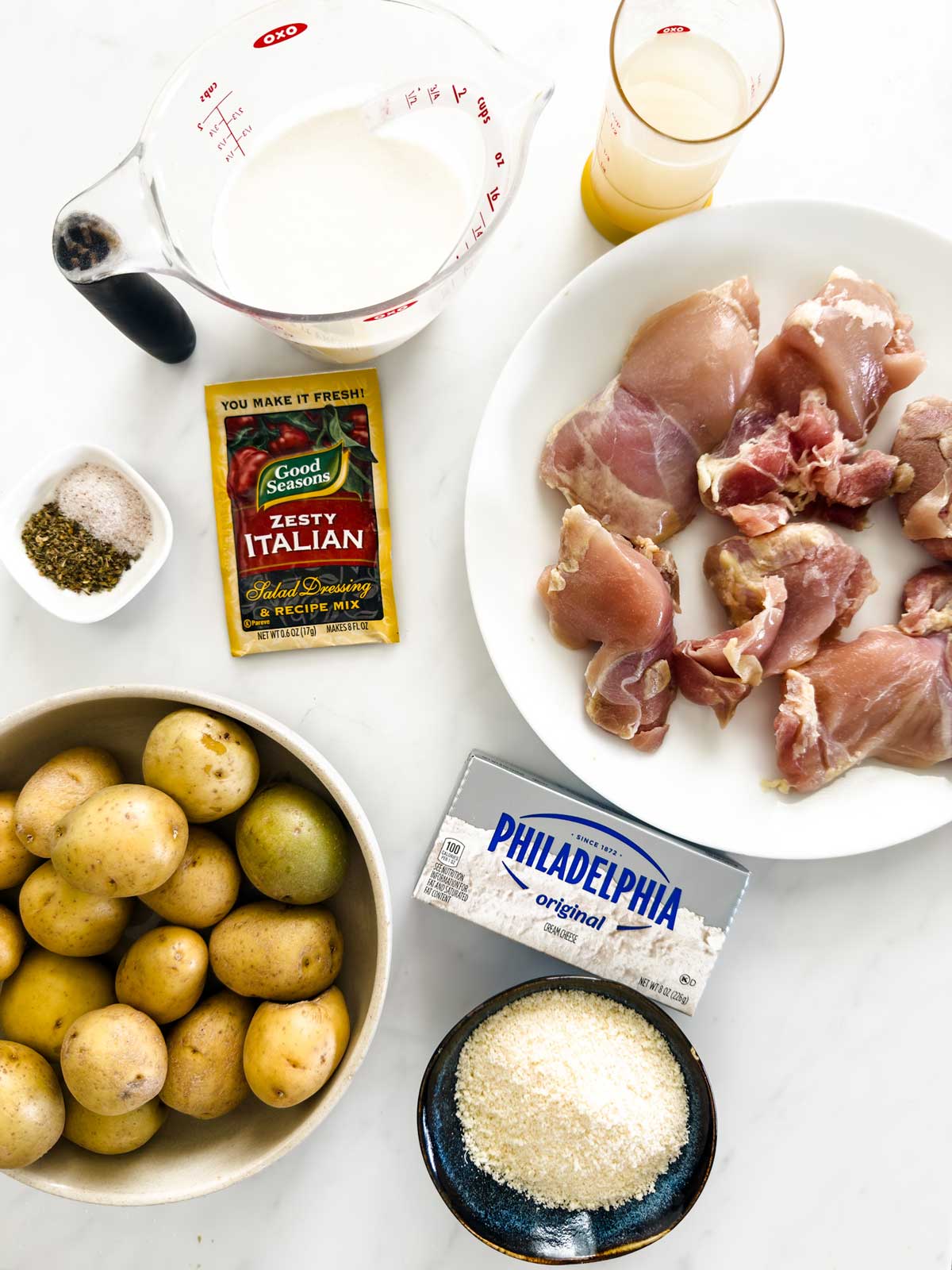 Ingredients for a recipe arranged on a white surface, including chicken thighs, baby potatoes, cream cheese, shredded parmesan, Italian dressing mix, herbs, milk, and a measuring cup with broth.