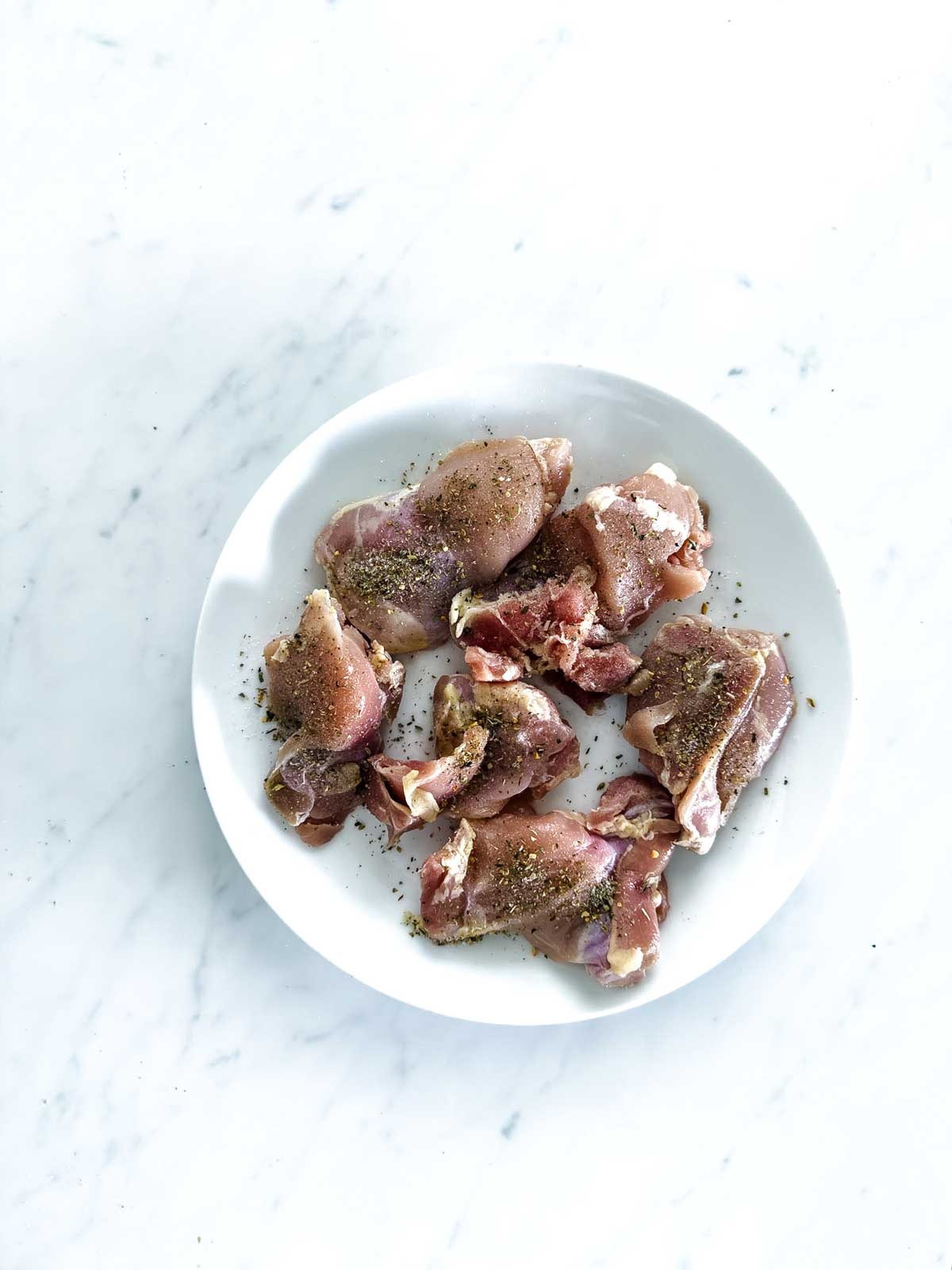 Raw, seasoned pieces of chicken on a white plate, placed on a white marble surface. The chicken is sprinkled with black pepper and other spices.