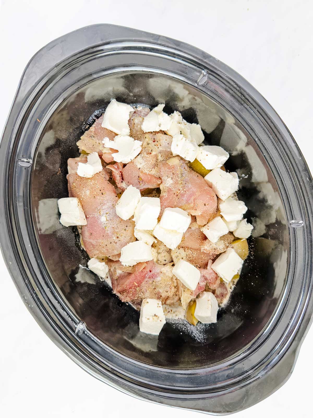 Raw chicken pieces seasoned with spices and topped with cubes of cream cheese are placed inside a black slow cooker on a white surface.