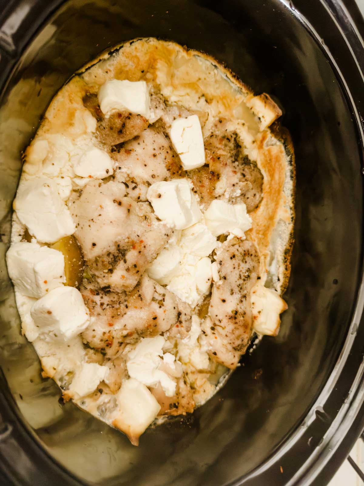 Chicken breasts cooked in a slow cooker, topped with melted cubes of cream cheese and sprinkled with seasoning. The dish appears creamy and flavorful.