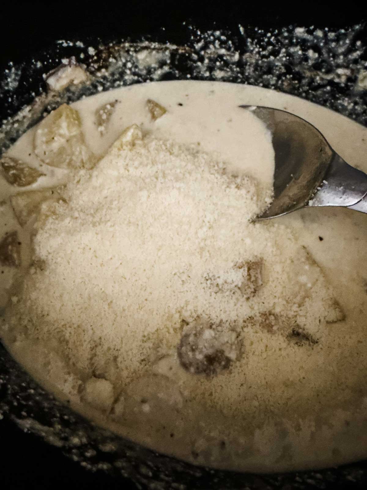 A close-up of a creamy dish with chunks of potato and a generous amount of grated parmesan cheese on top, being stirred with a metal spoon.