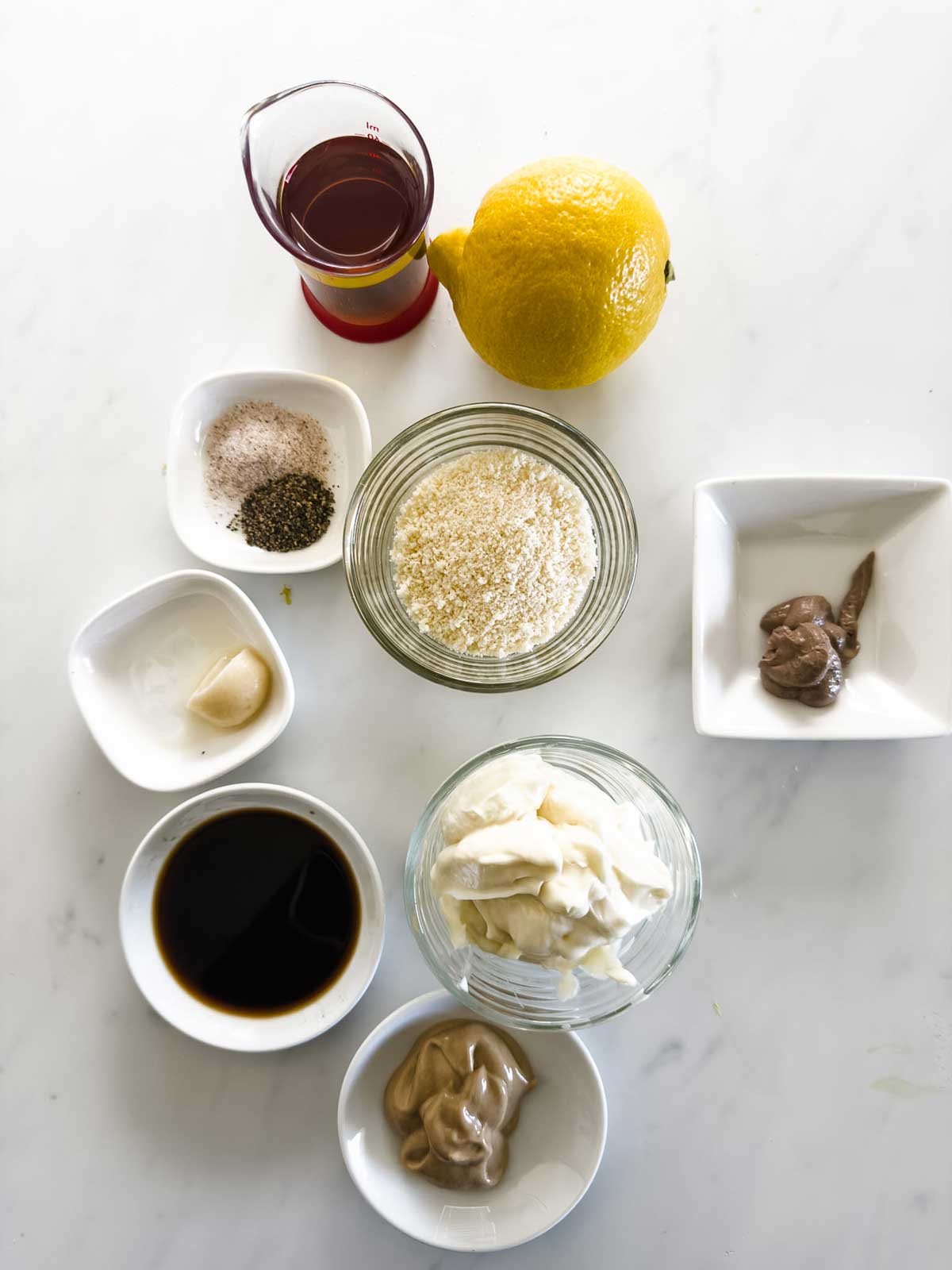 Overhead view of various ingredients in small bowls on a white surface: oil in a glass, a lemon, spices, a garlic clove, parmesan, mayonnaise, Dijon mustard, Worcestershire sauce, and anchovy paste.