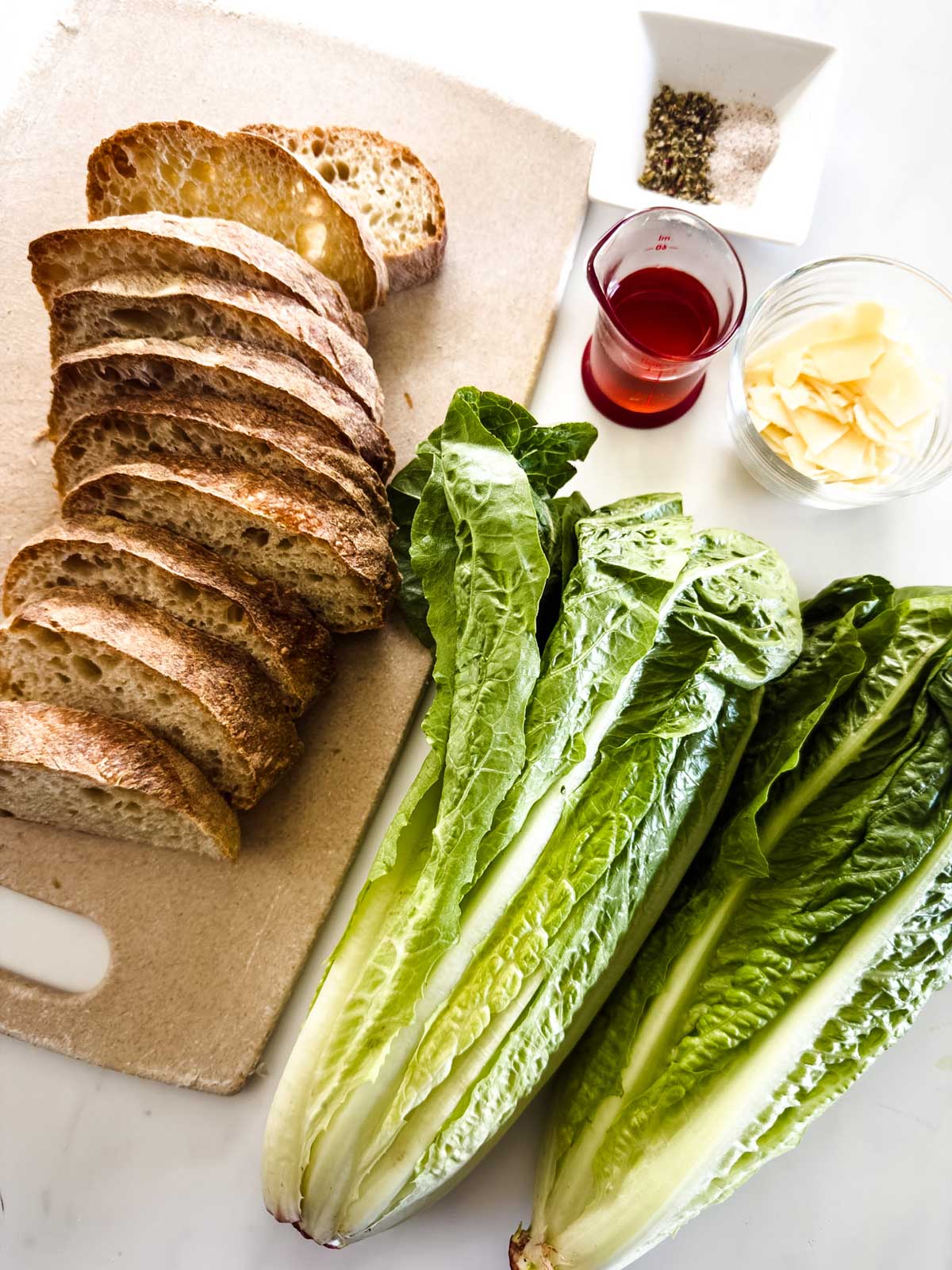 Sliced bread, two heads of romaine lettuce, a small glass of red liquid, a bowl of Parmesan slices, and a dish of salt and pepper are arranged on a cutting board and a white surface.