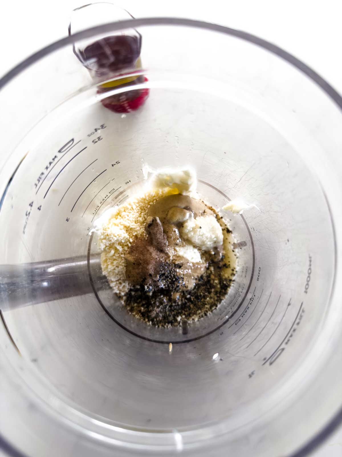 A top-down view of a glass measuring cup containing yogurt, spices, and seasonings, ready to be mixed, with a metal immersion blender inside the cup.
