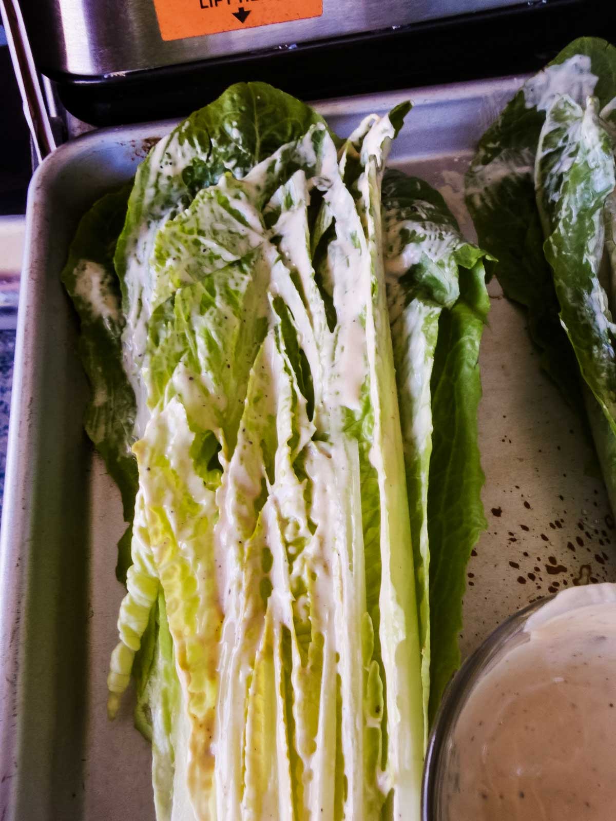 A close-up of romaine lettuce leaves spread with creamy dressing on a metal tray, with a small bowl of extra dressing nearby.