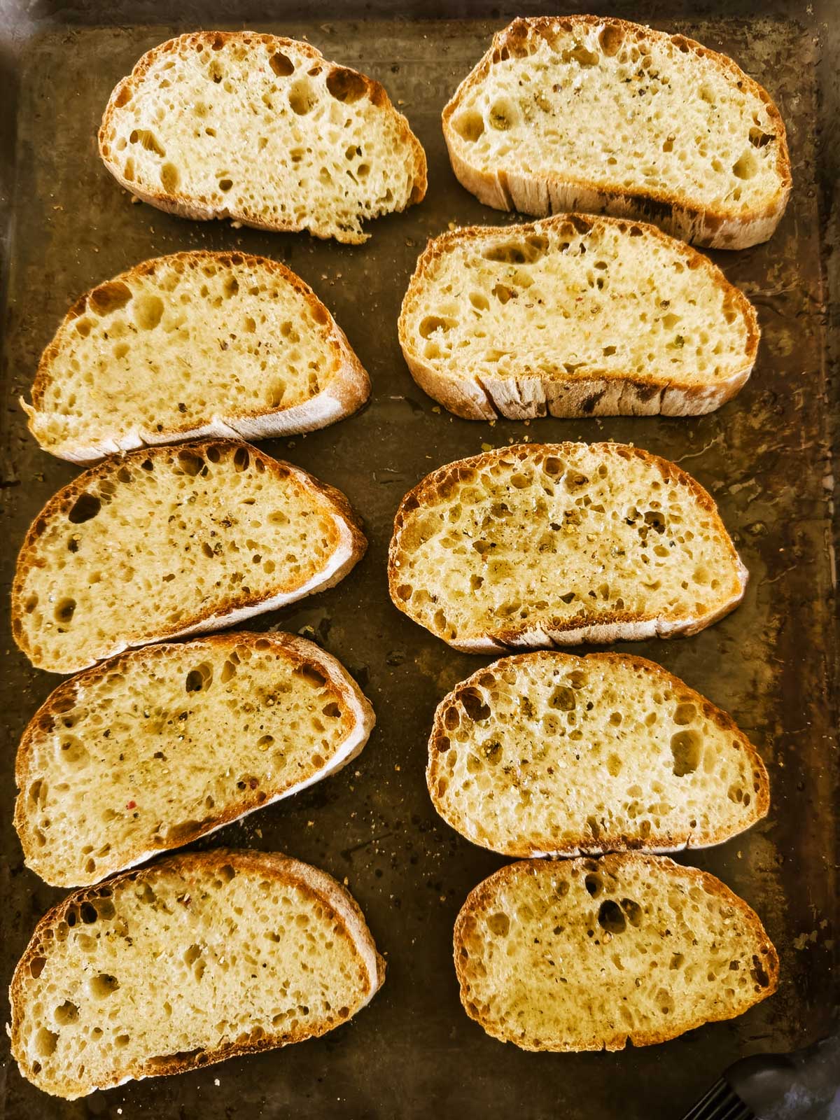 Ten slices of rustic bread are arranged in two columns on a metal baking sheet. The bread has a golden, crispy crust and an airy, open crumb with visible holes.